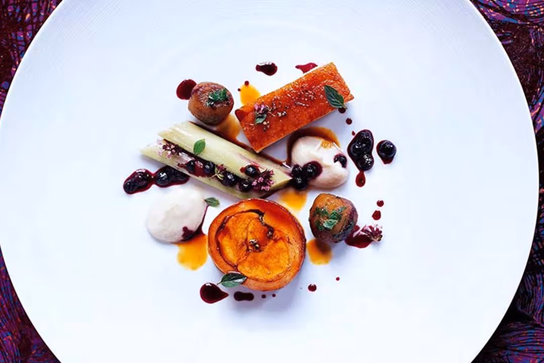 Gourmet dish with seared meat, grilled vegetables, and berry sauce artfully plated on a large white plate.