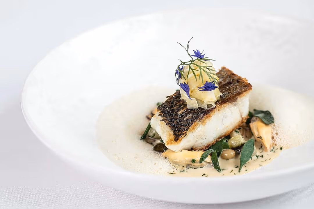 Pan-seared fish fillet with crispy skin, garnished with edible flowers and herbs, served with creamy sauce and vegetables in a white bowl.