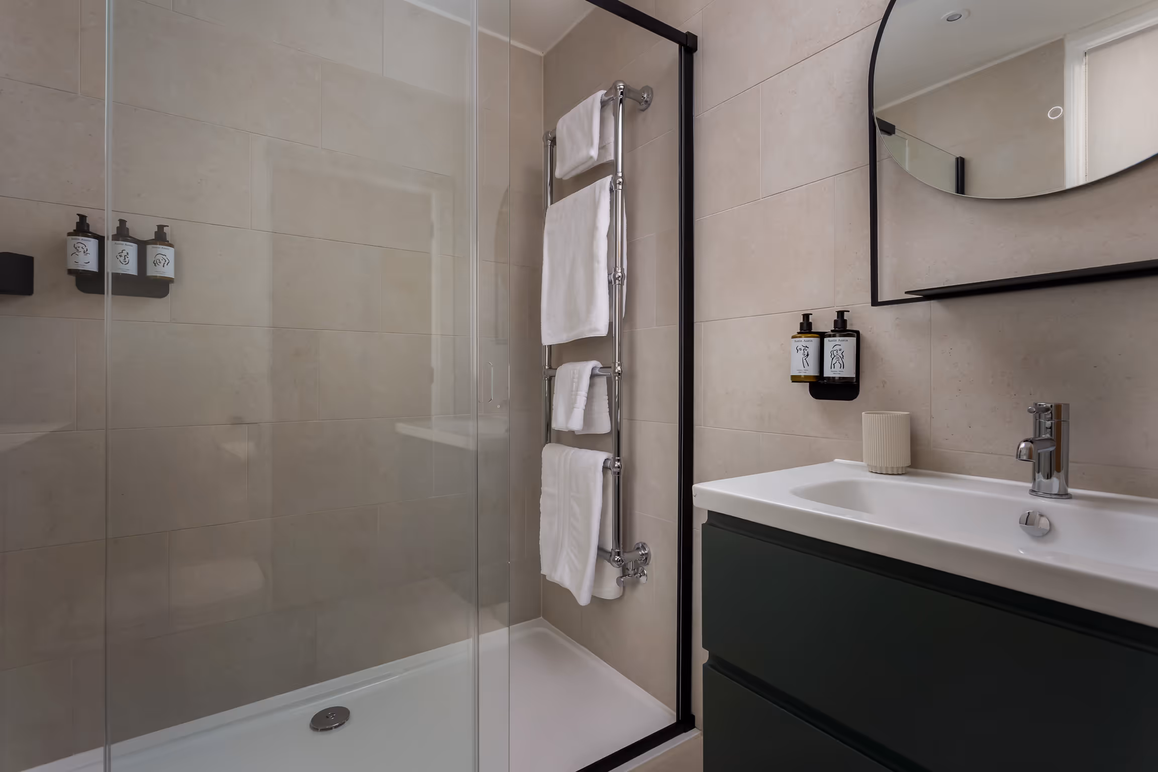 Modern bathroom with beige tiled walls, glass shower enclosure, chrome towel rack with white towels, and a sink with dark green cabinet and round mirror.