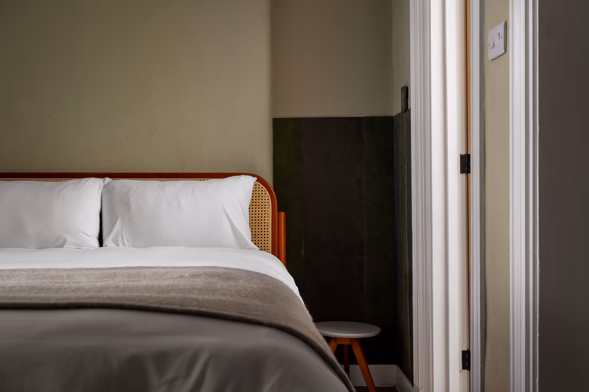 Side view of a neatly made bed with white pillows, beige blanket, and a rattan headboard next to a small round white stool near a doorway.