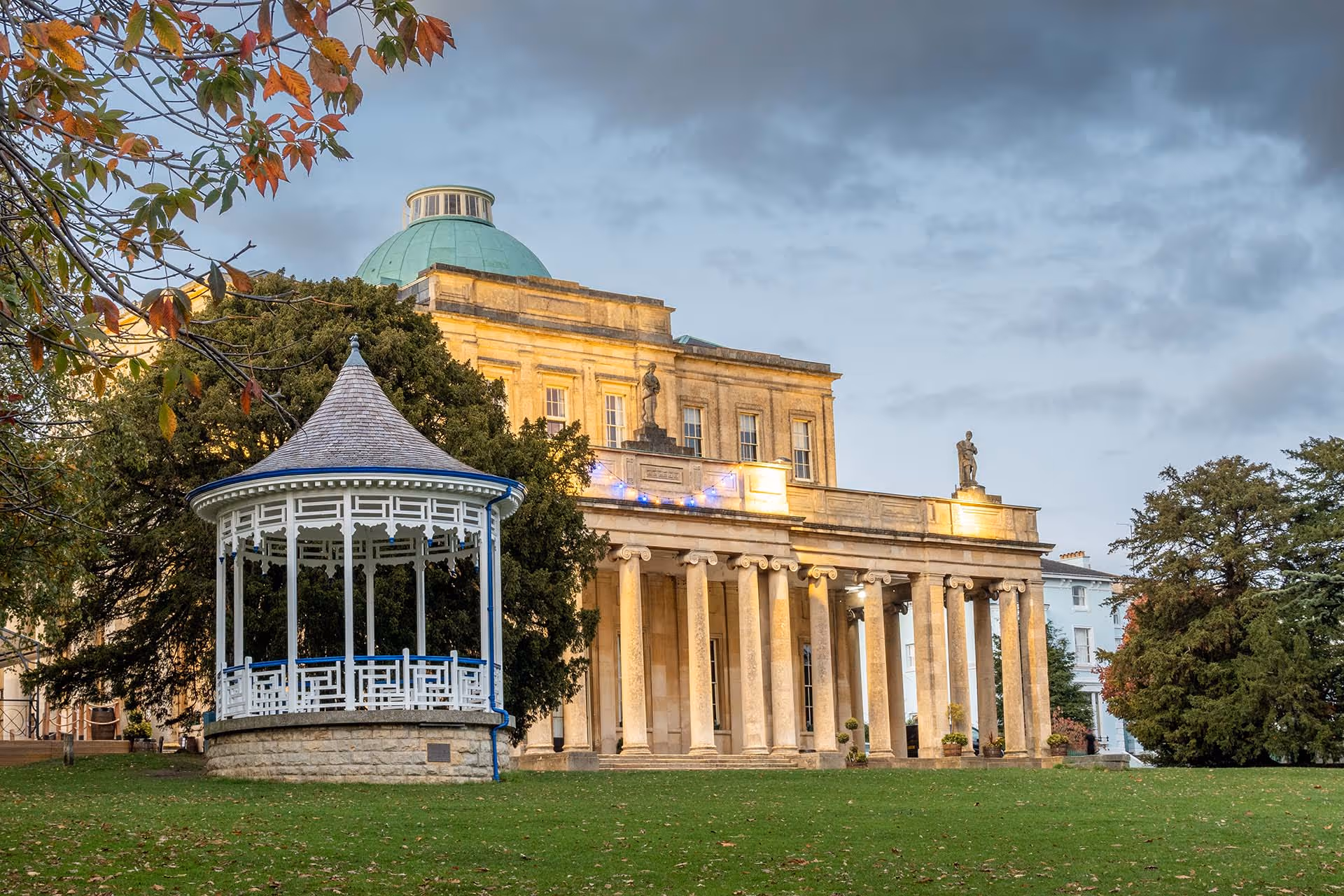 Historic neoclassical building with tall columns and statues on the roof, next to a white gazebo on a green lawn under a cloudy sky.