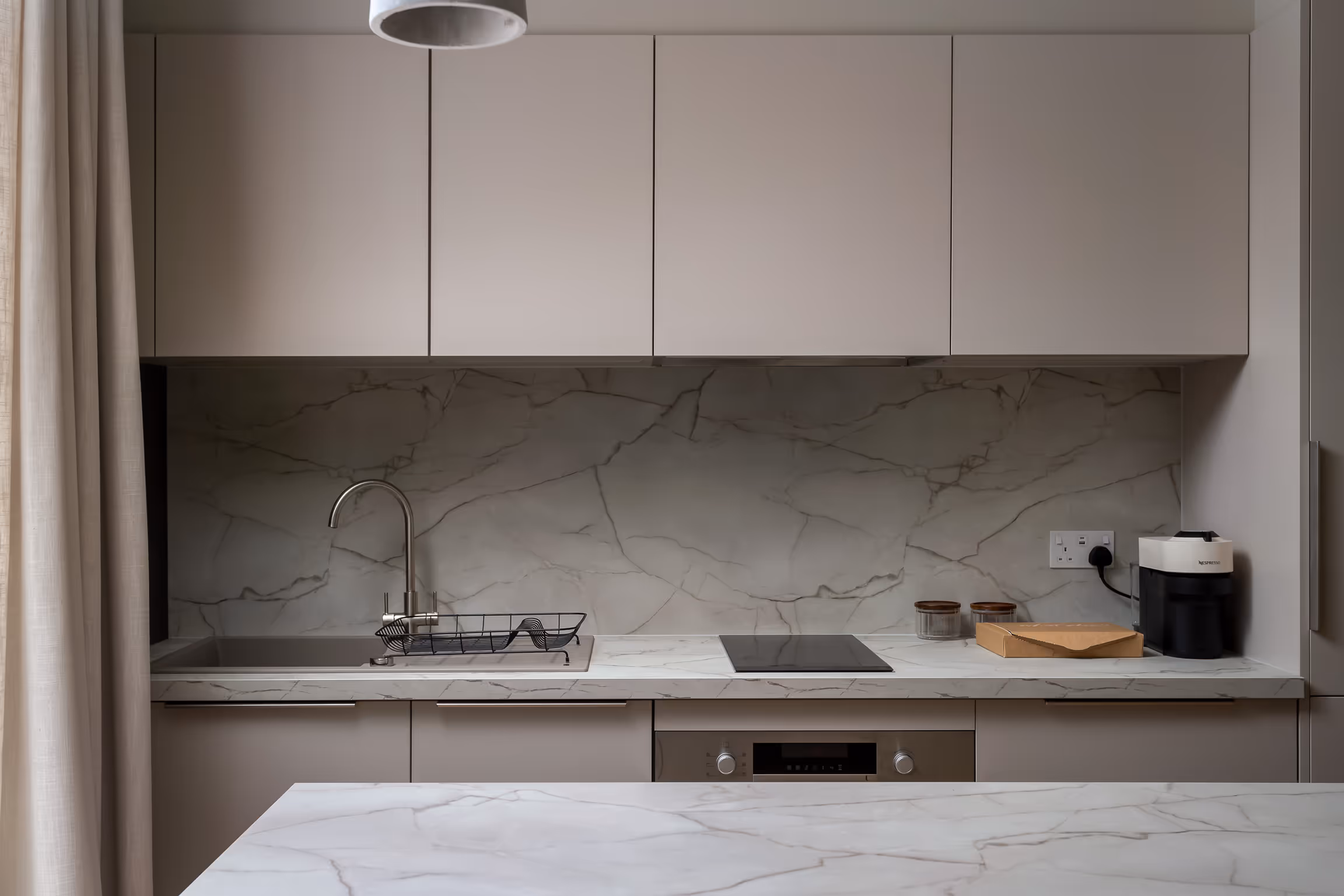 Minimalist kitchen with beige cabinets, marble countertops, sink with drying rack, electric stovetop, and coffee machine.