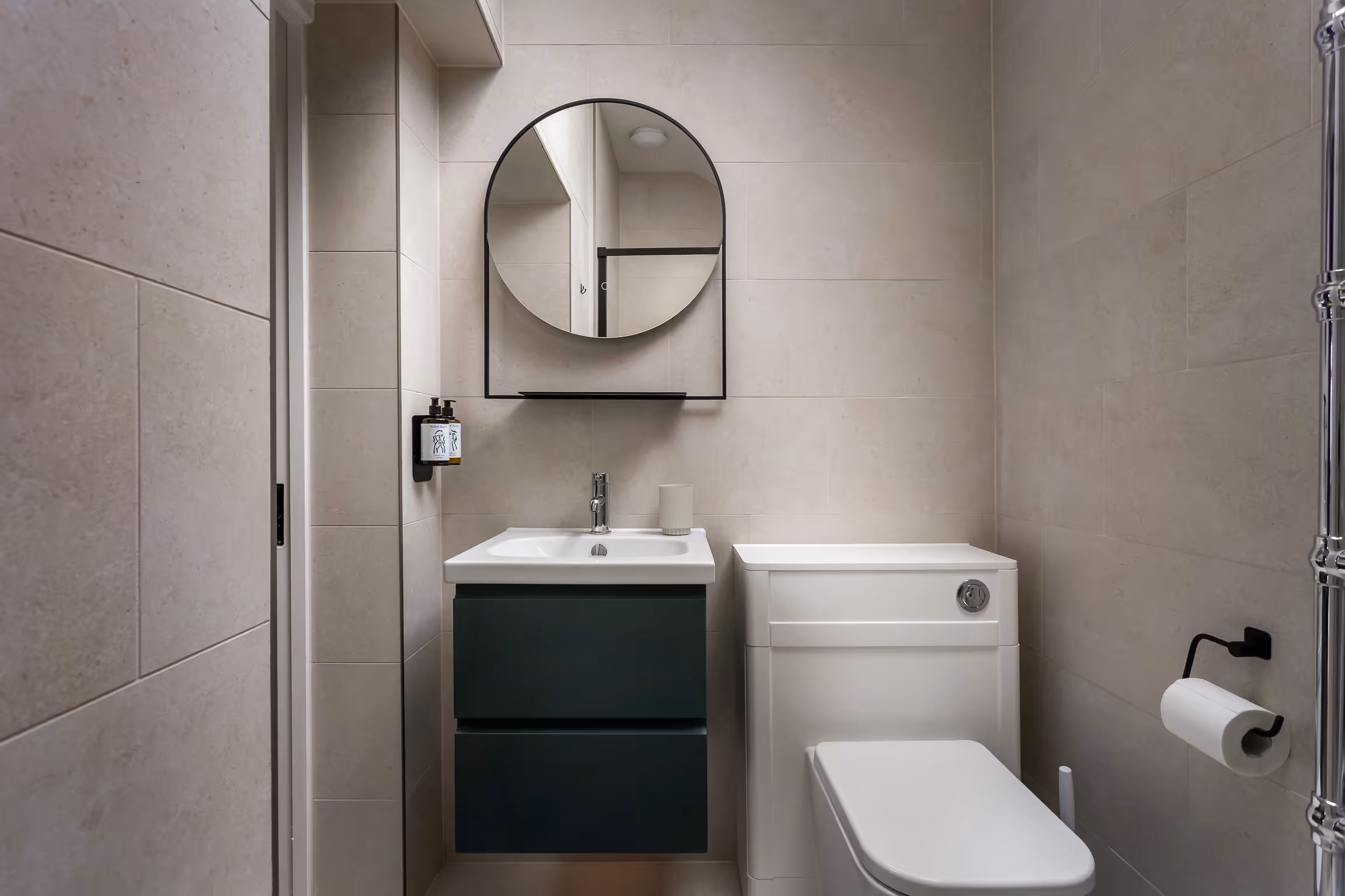 Compact modern bathroom with a white toilet, dark green vanity with sink, round mirror, and beige tiled walls.