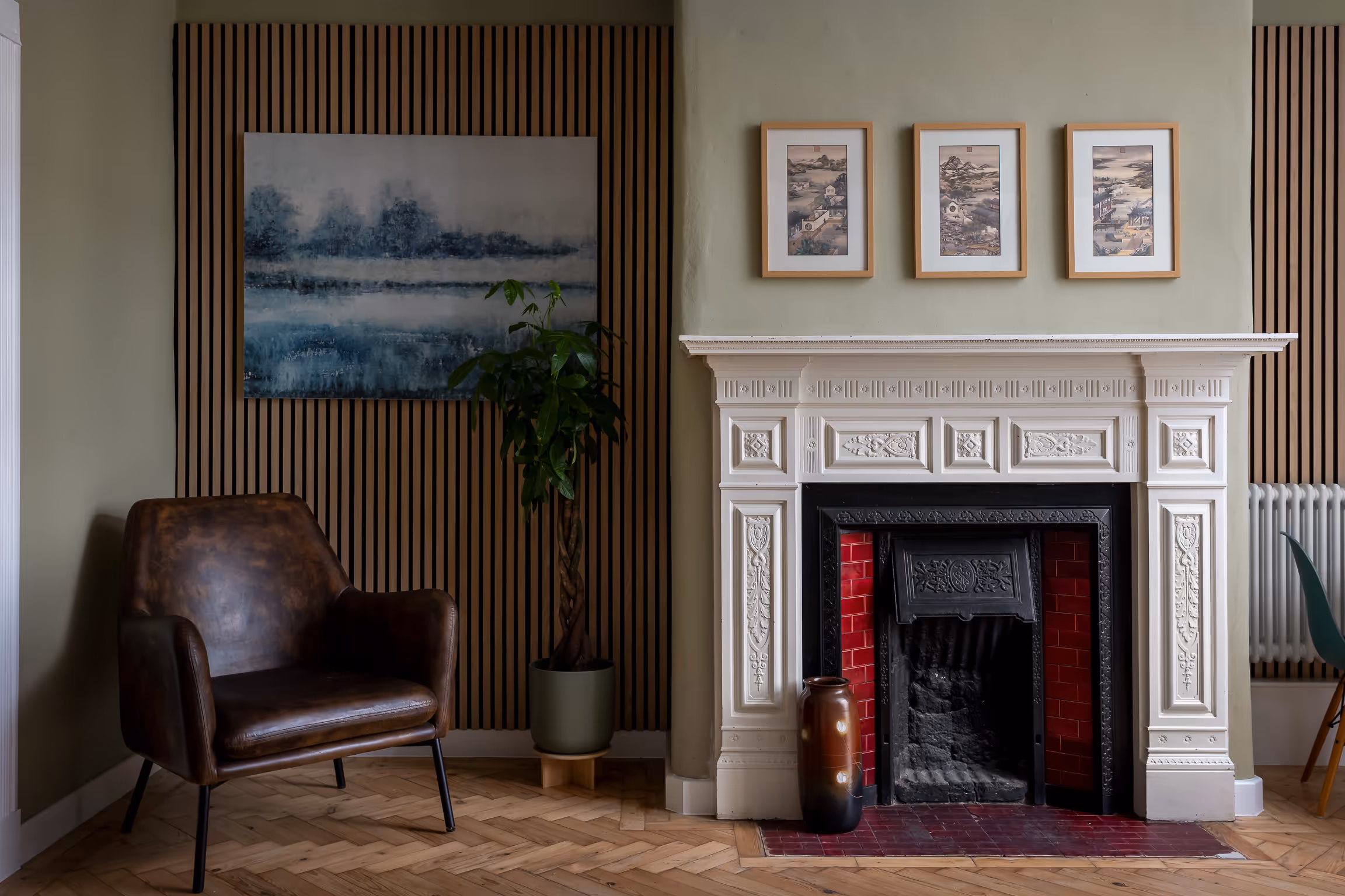Cozy corner with a brown leather armchair, potted plant, white ornate fireplace with red tiles, and framed landscape art on walls.