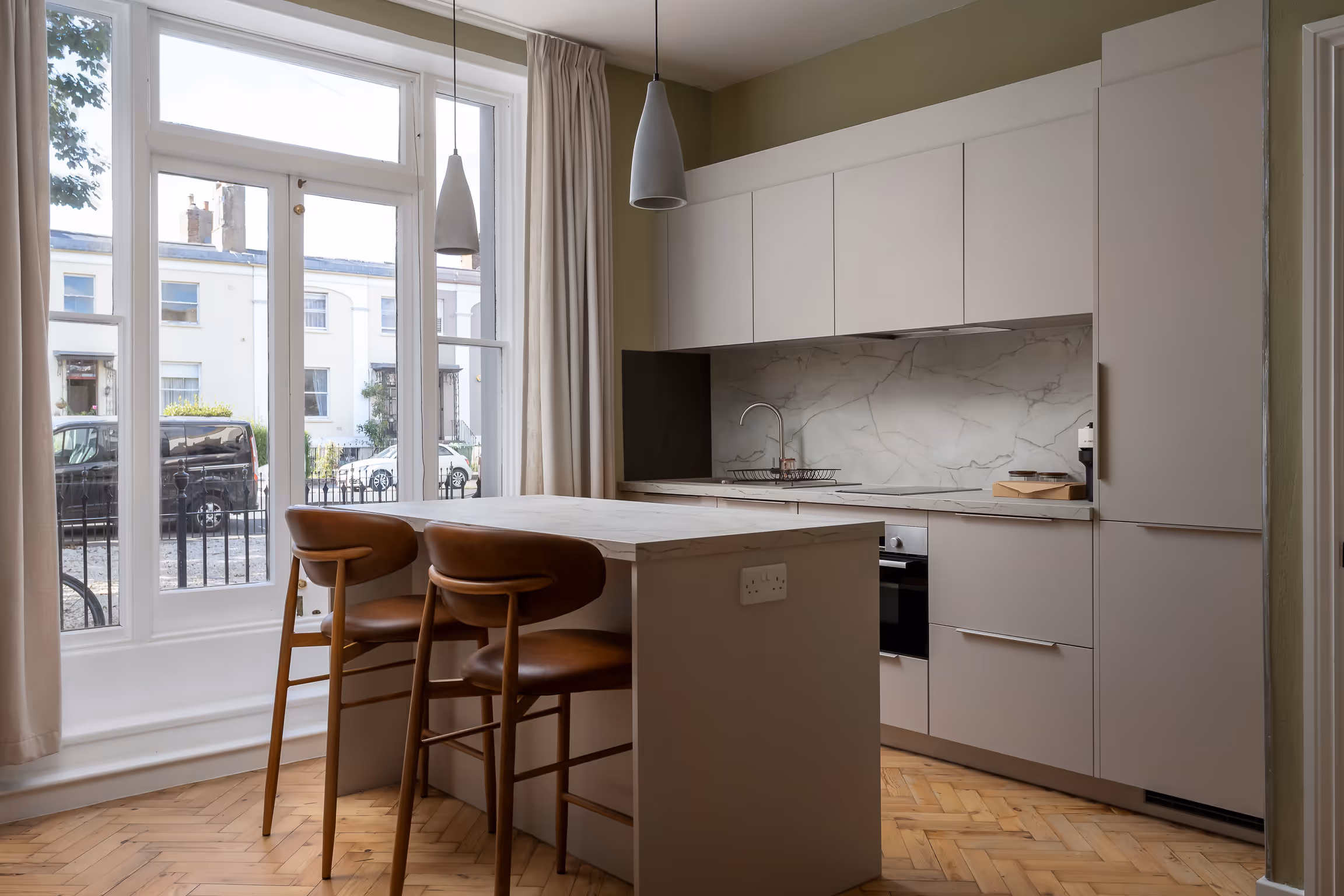 Modern kitchen with marble countertop island, two brown leather chairs, and large windows with beige curtains.