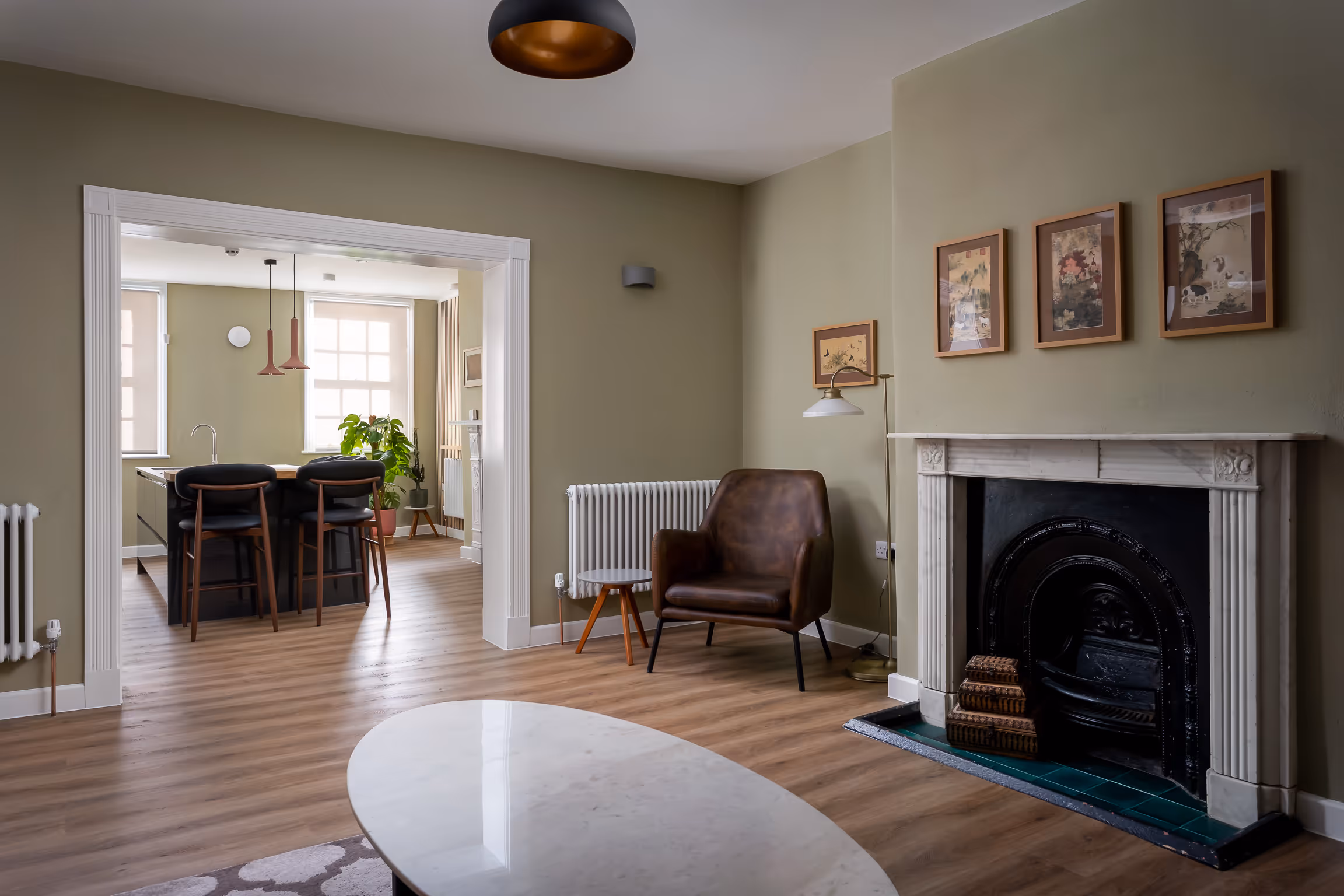 Living room with brown leather armchair, marble coffee table, decorative fireplace, and adjoining kitchen with bar stools and pendant lights.