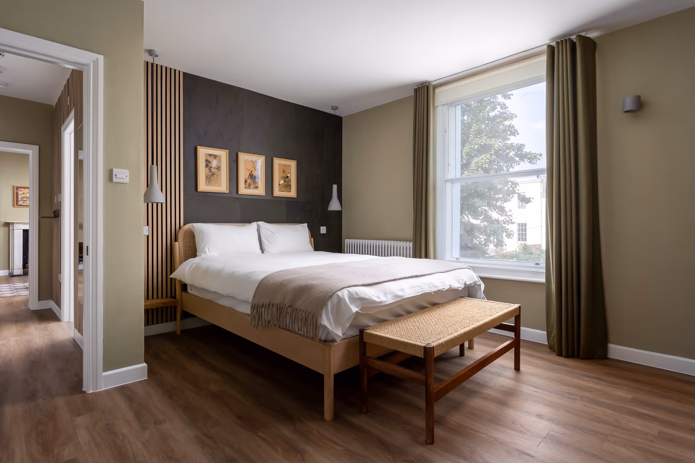 Modern bedroom with a wooden bed, white bedding, beige throw, three framed artworks above the headboard, large window with green curtains, and a woven bench at the foot of the bed.