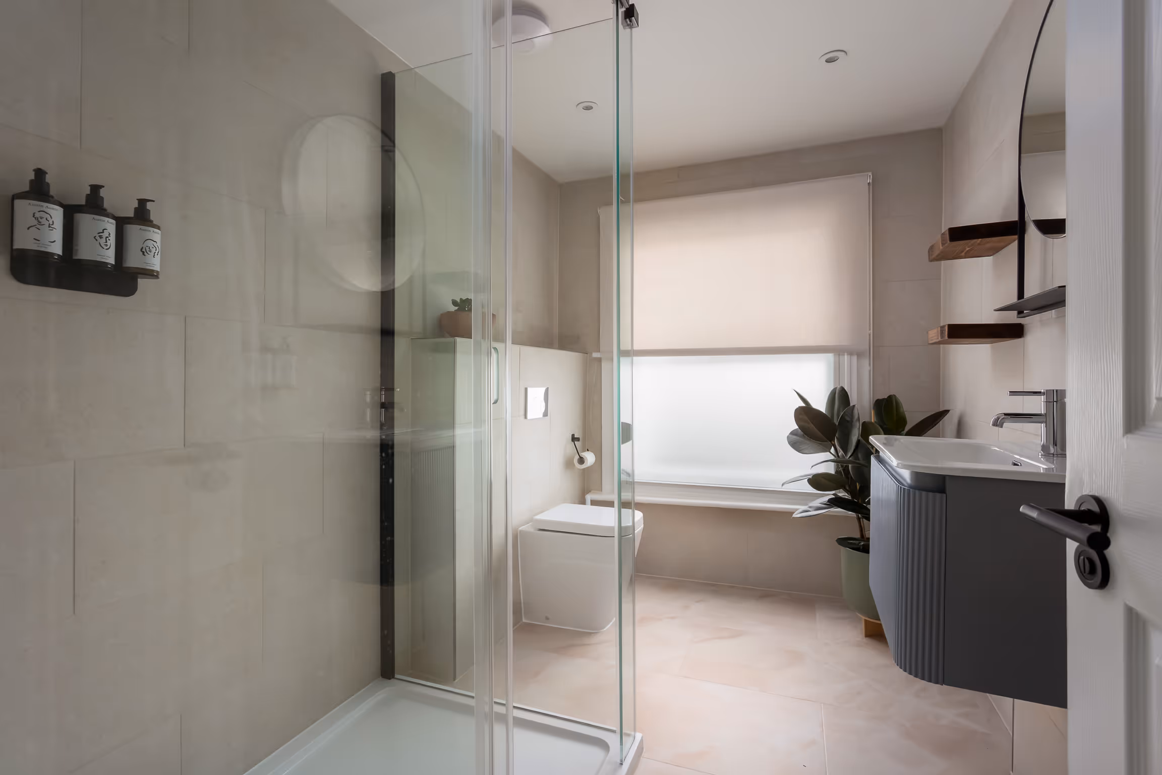 Modern bathroom with walk-in glass shower, wall-mounted toilet, floating sink vanity, and a large window with a beige roller shade.