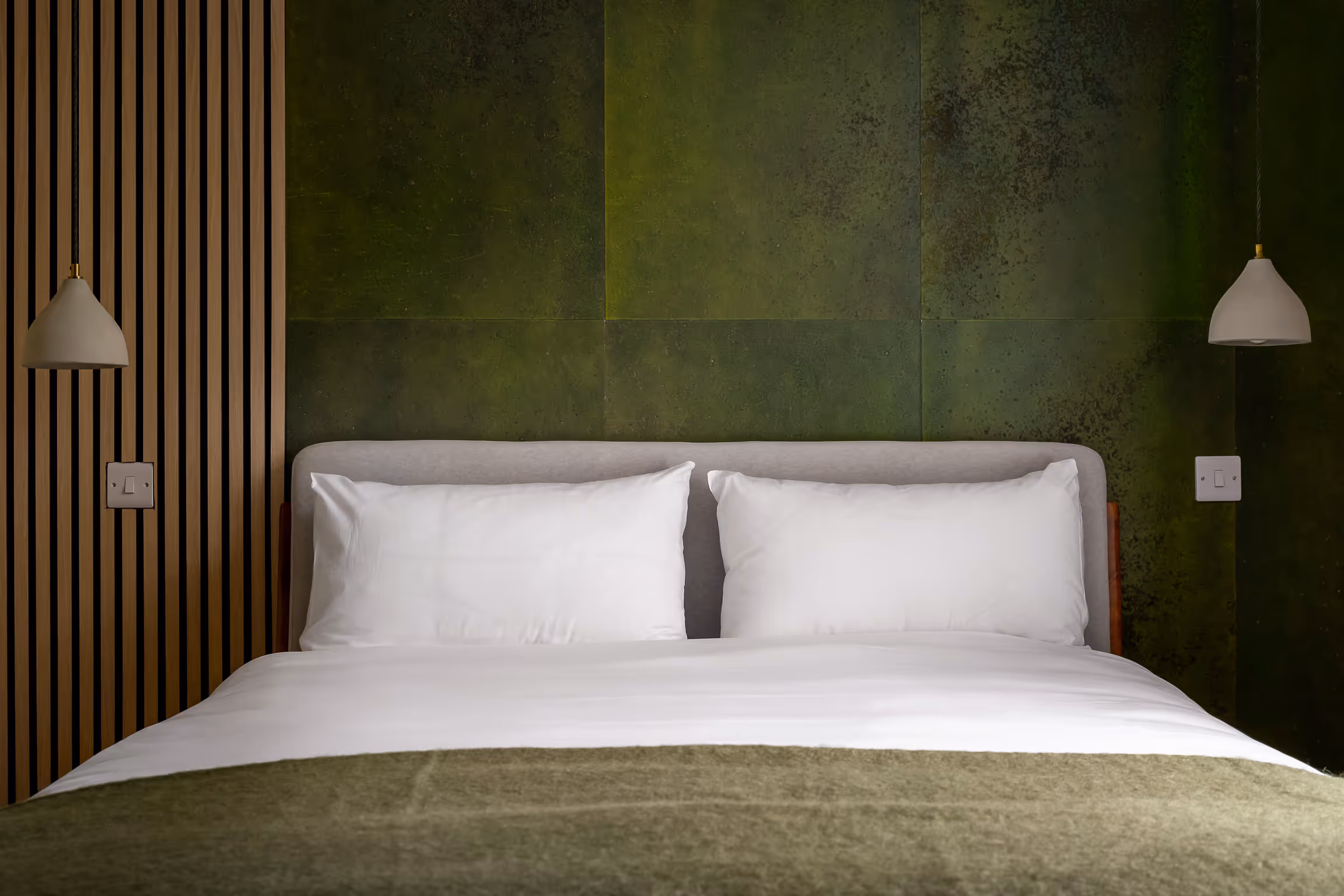 Neatly made bed with white pillows and sheets, green blanket, and green textured wall behind with two hanging pendant lights.