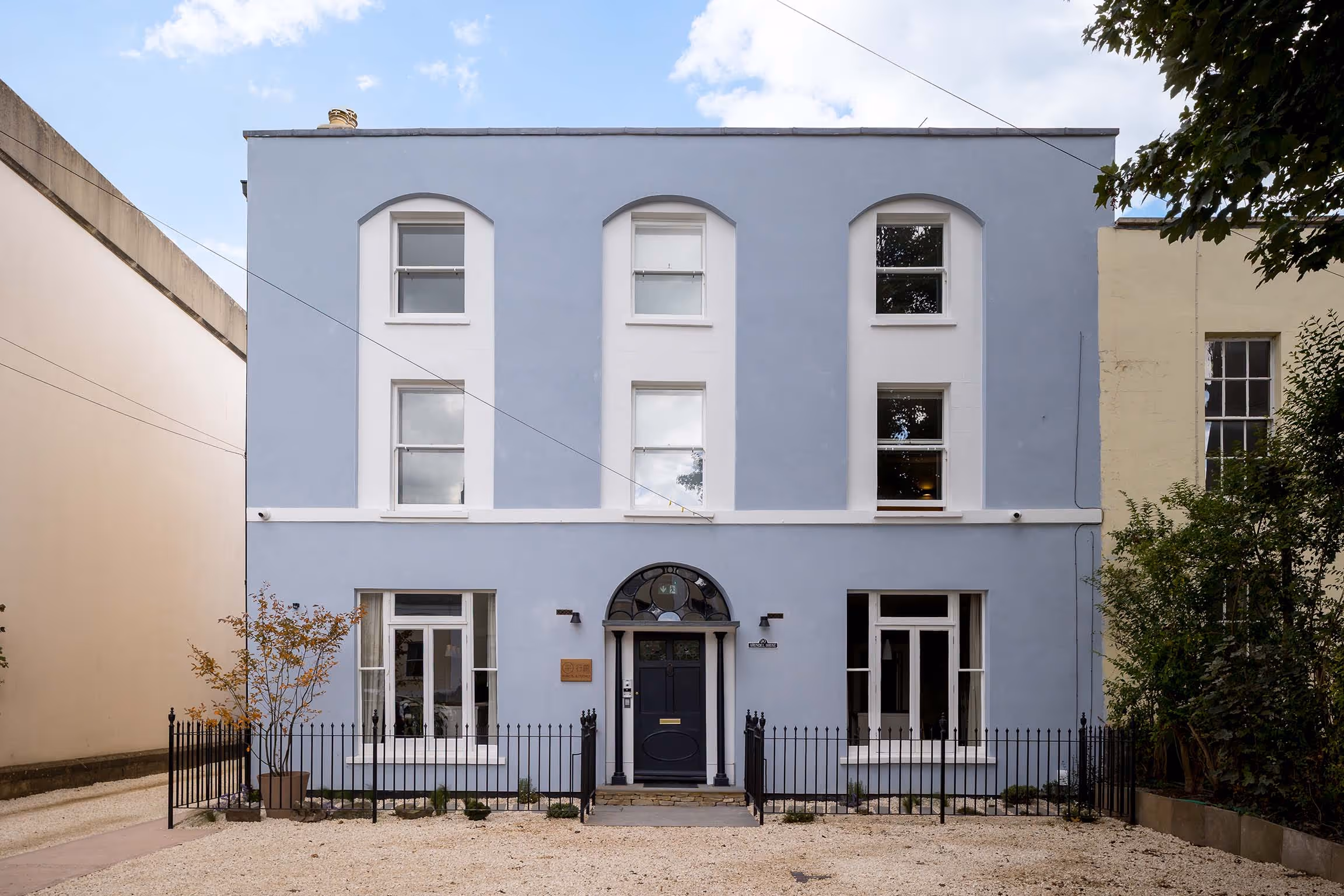 Two-story light blue building with white window frames, black front door with an arched transom window, surrounded by a black metal fence and small plants.