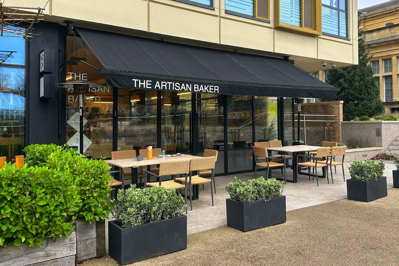 Outdoor seating area of The Artisan Baker café with tables, wicker chairs, and planter boxes with greenery.