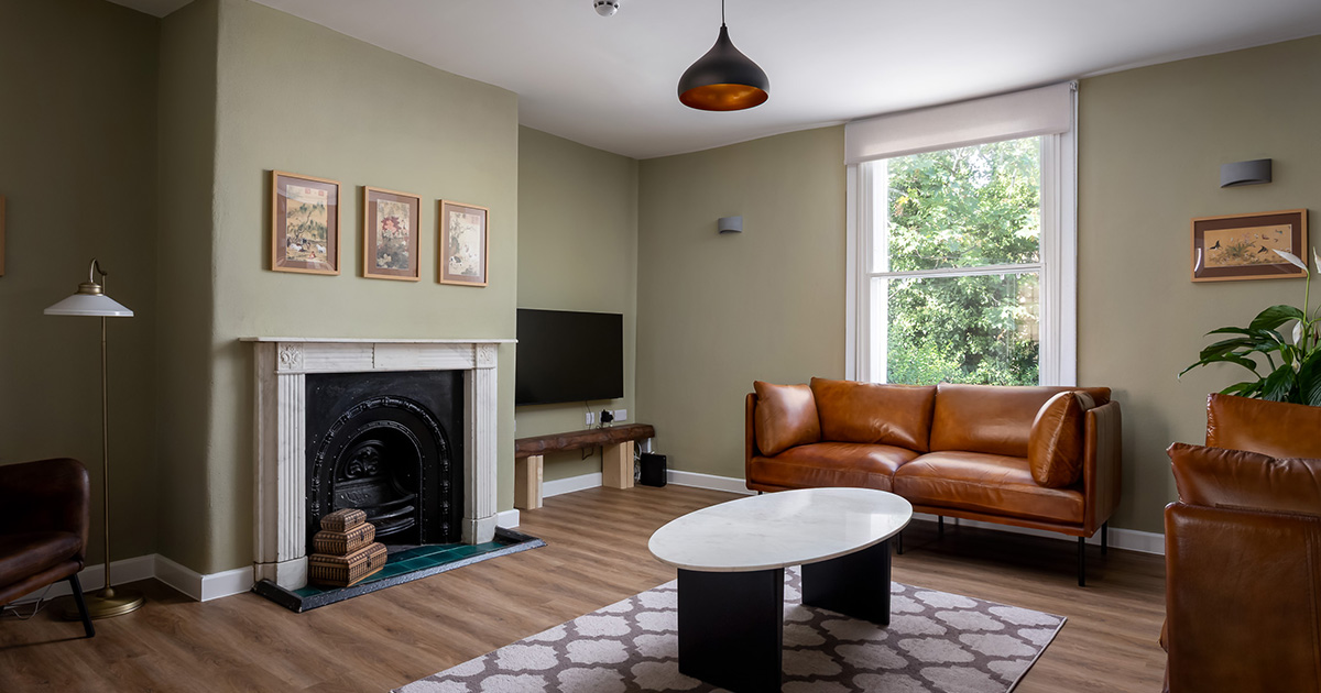 Modern living room with a brown leather sofa and chairs, a white marble coffee table on a patterned rug, wooden floor, green walls, and a white-framed window showing trees outside.