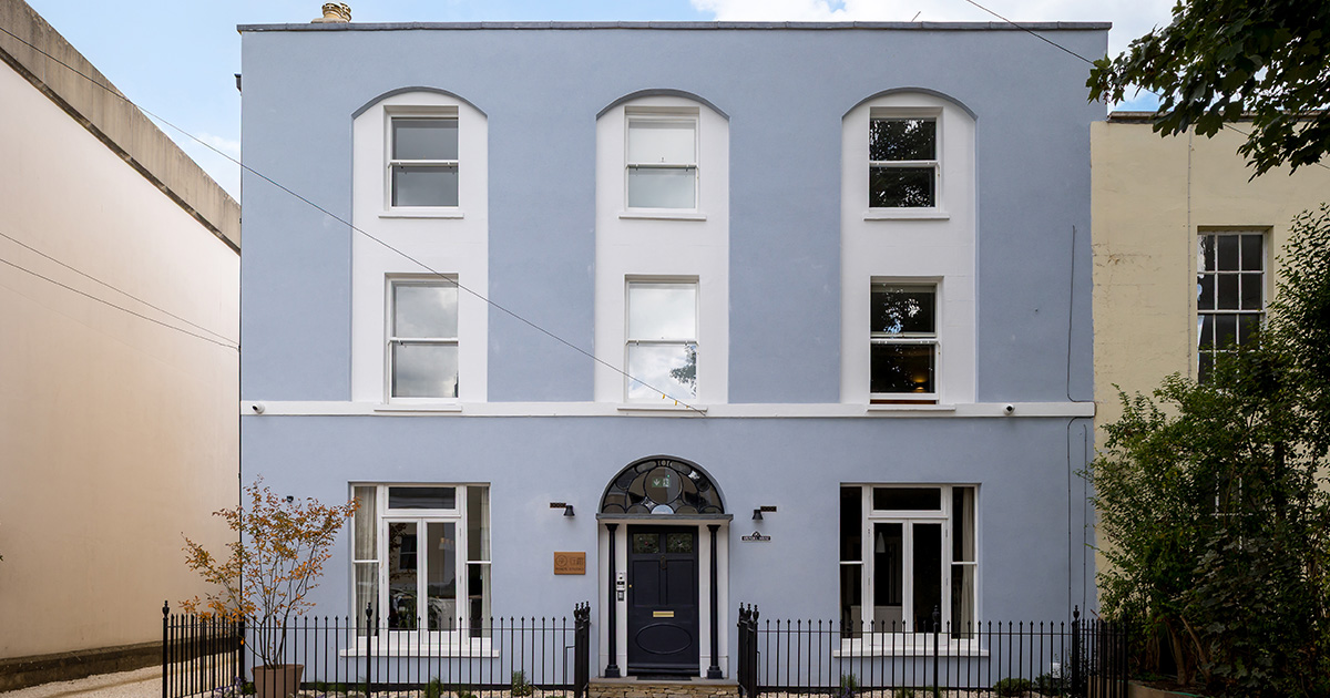 Front view of a two-story light blue building with arched windows, a black door with decorative black columns, and a black iron fence in front.