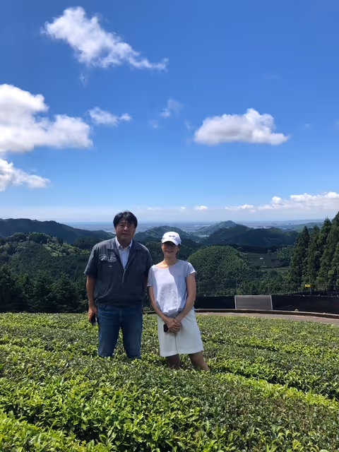 A man and woman standing in a green tea field with mountains and a blue sky with clouds in the background.
