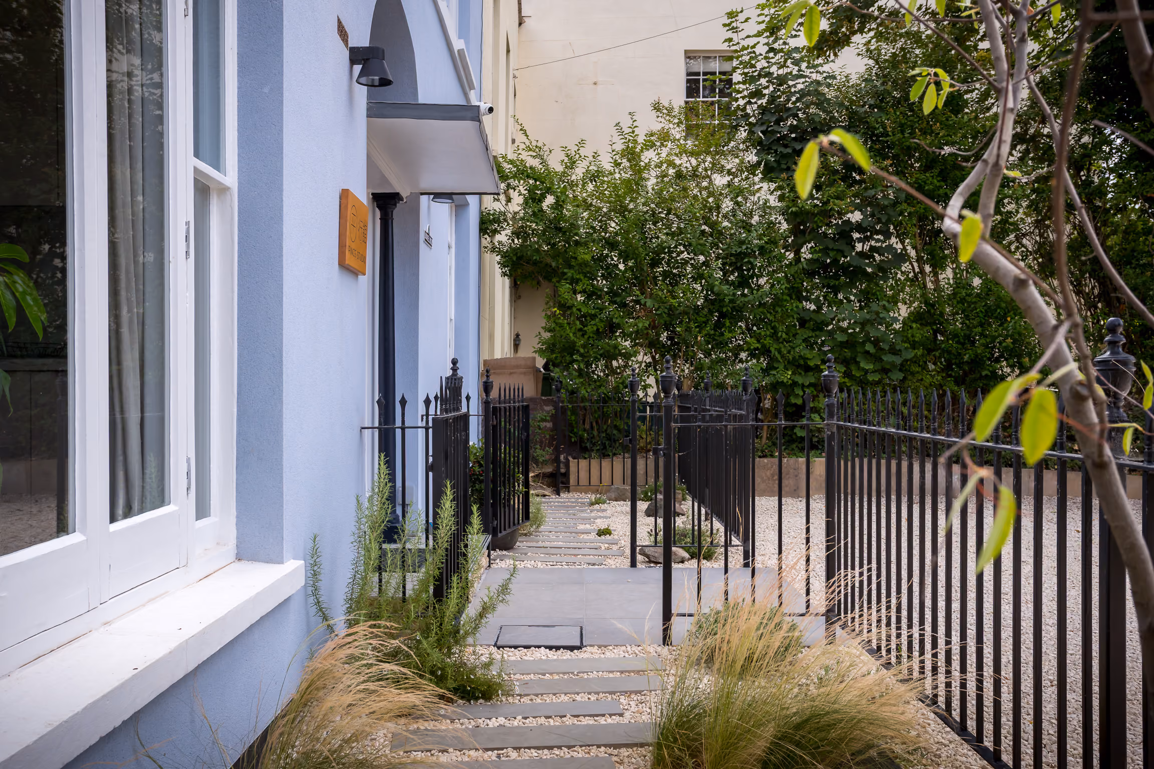 Blue house exterior with black iron fence, stone path, greenery, and trees in a garden area.