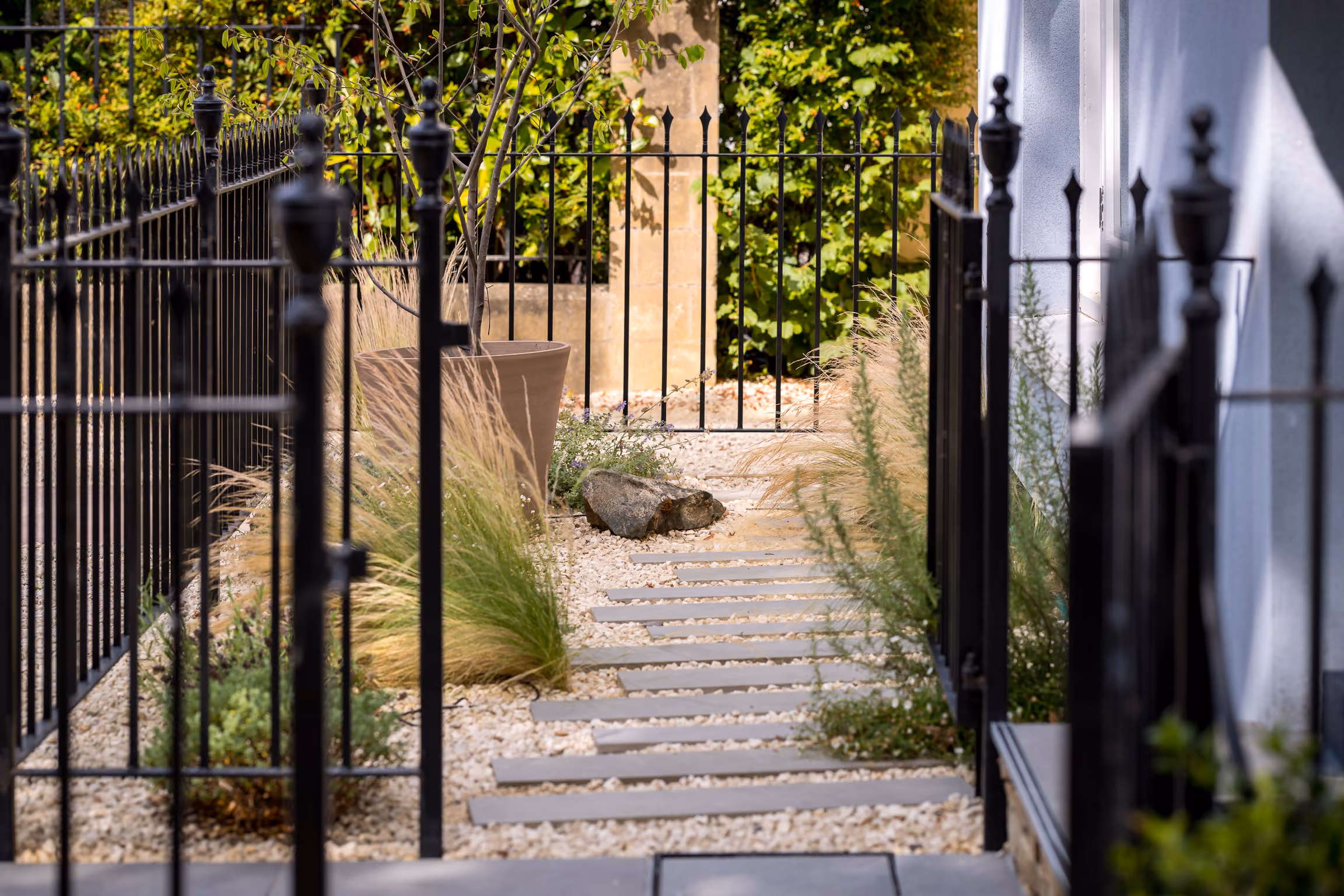 Garden path with stepping stones, gravel, ornamental grasses, and black metal fencing on both sides.