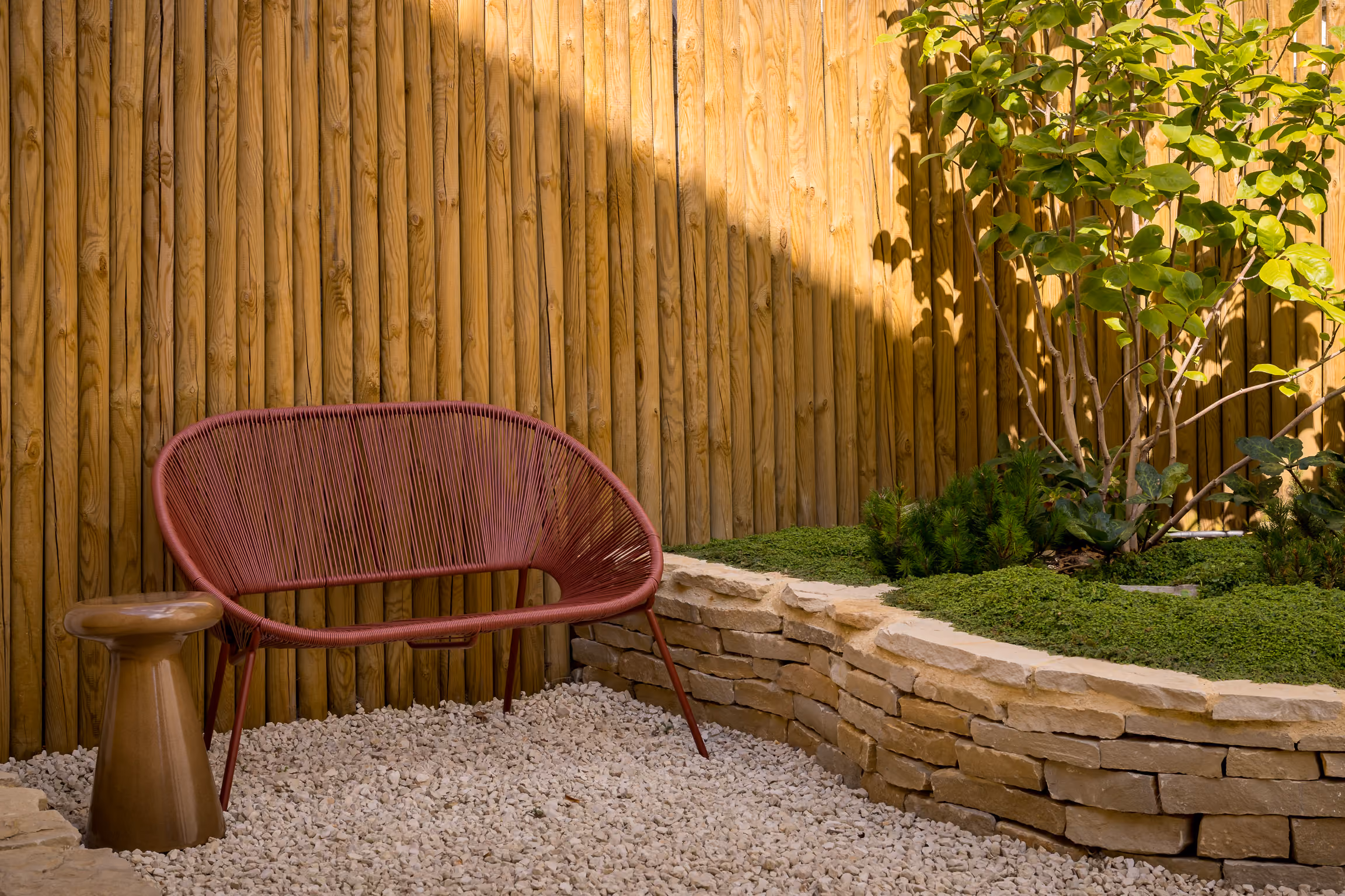 Red wire outdoor chair and small wooden stool on gravel next to stone planter with green plants.