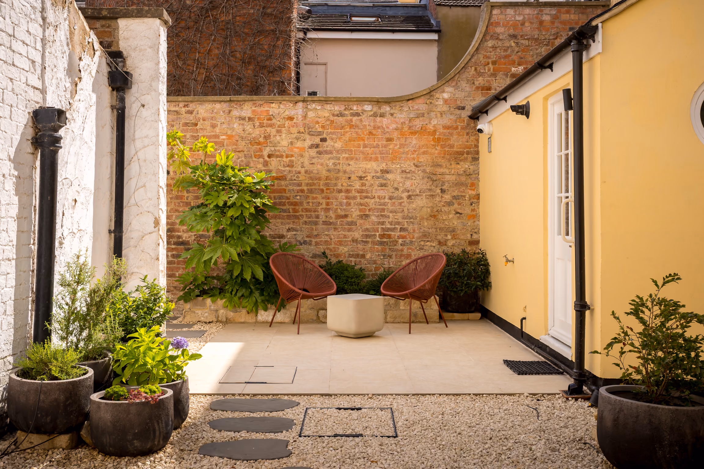 Small sunny courtyard with two red chairs, a square table, potted plants, and brick and yellow walls.