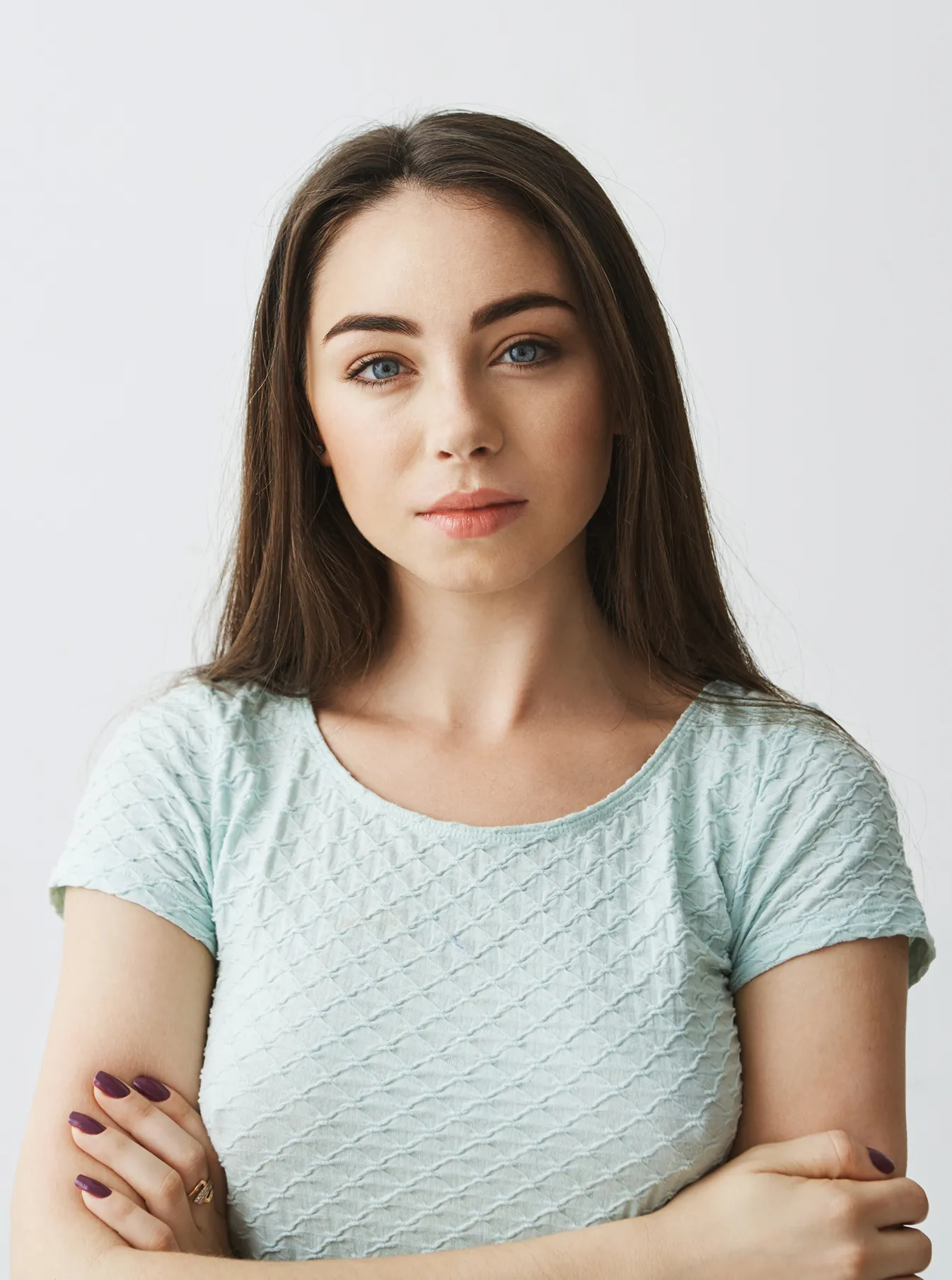 Portrait of a serious young woman with long brown hair and blue eyes wearing a light blue textured shirt, arms crossed.