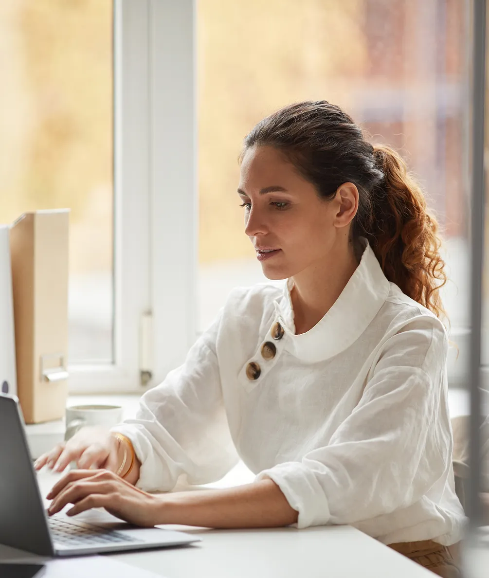 Woman with curly hair tied back typing on a laptop at a bright desk near a window.