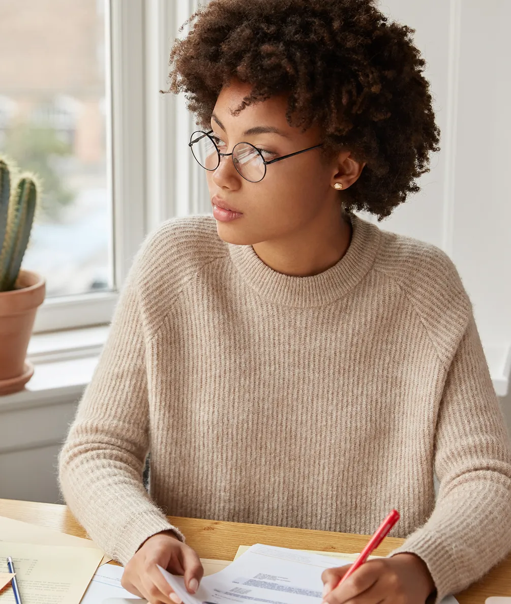 Woman with curly hair and glasses wearing a beige sweater, writing with a red pen at a desk near a window.