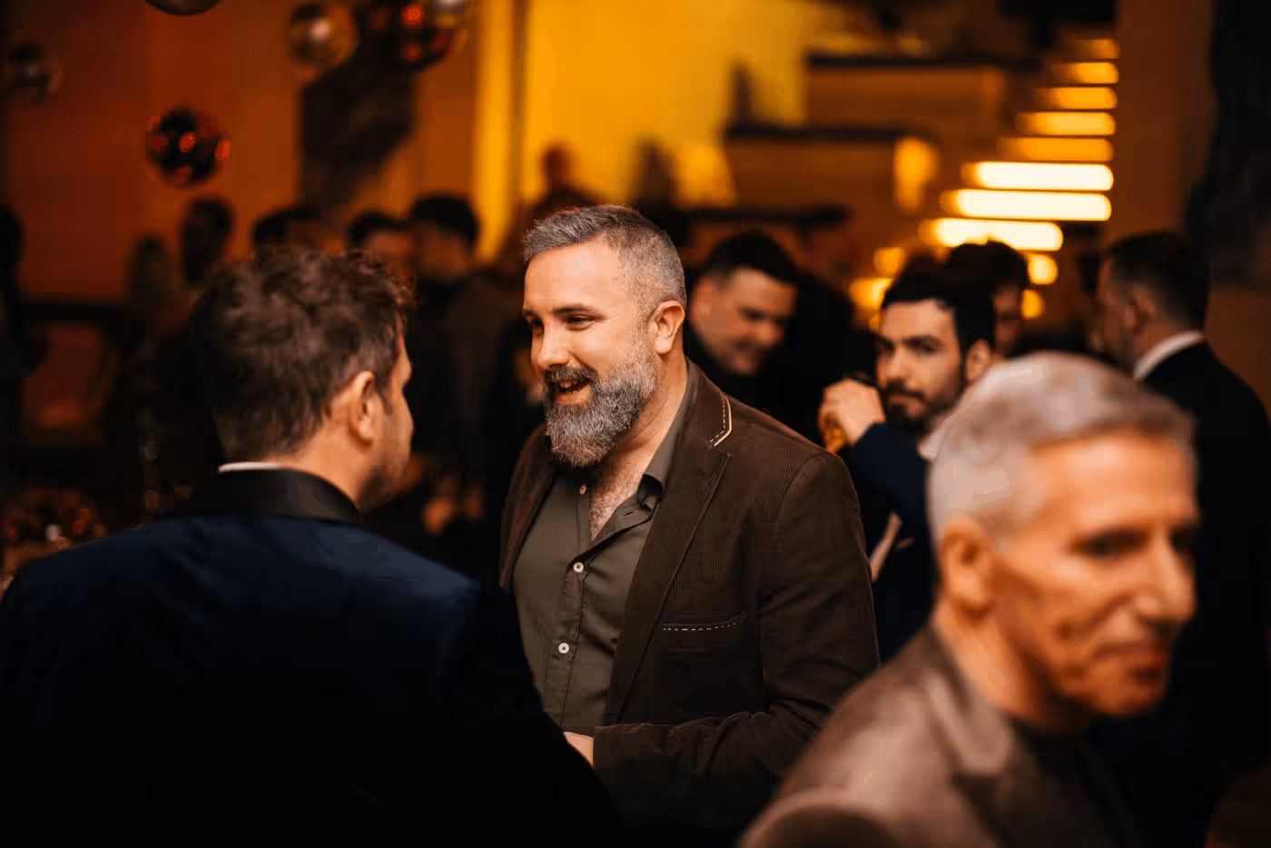 Man with gray beard and brown jacket smiling and talking to another man at a social event with warm lighting and blurred background of guests and stairs.