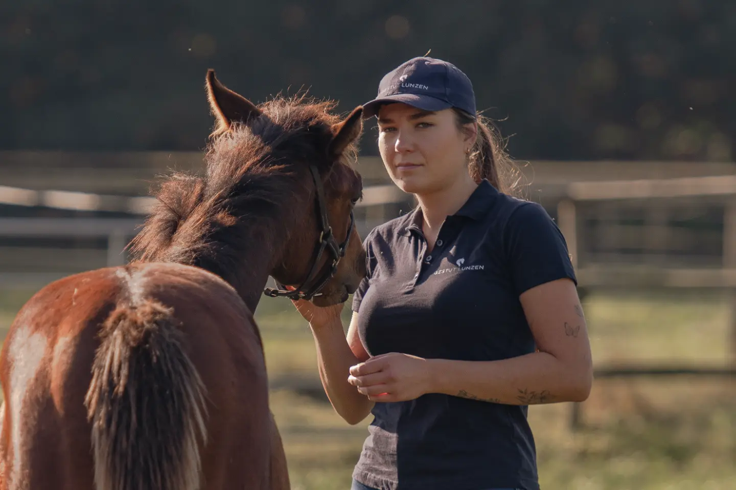 Frau in dunklem Polohemd und Mütze steht neben braunem Pferd auf einer Weide.