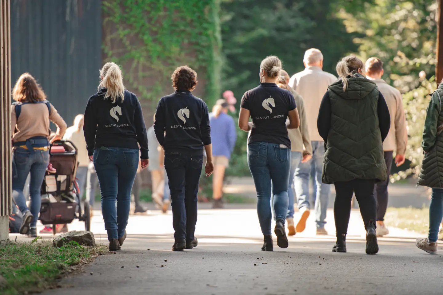 Mehrere Personen in Freizeitkleidung gehen auf einem sonnigen Weg, einige tragen schwarze Pullover mit dem Aufdruck ‚Gestüt Lunzen‘.