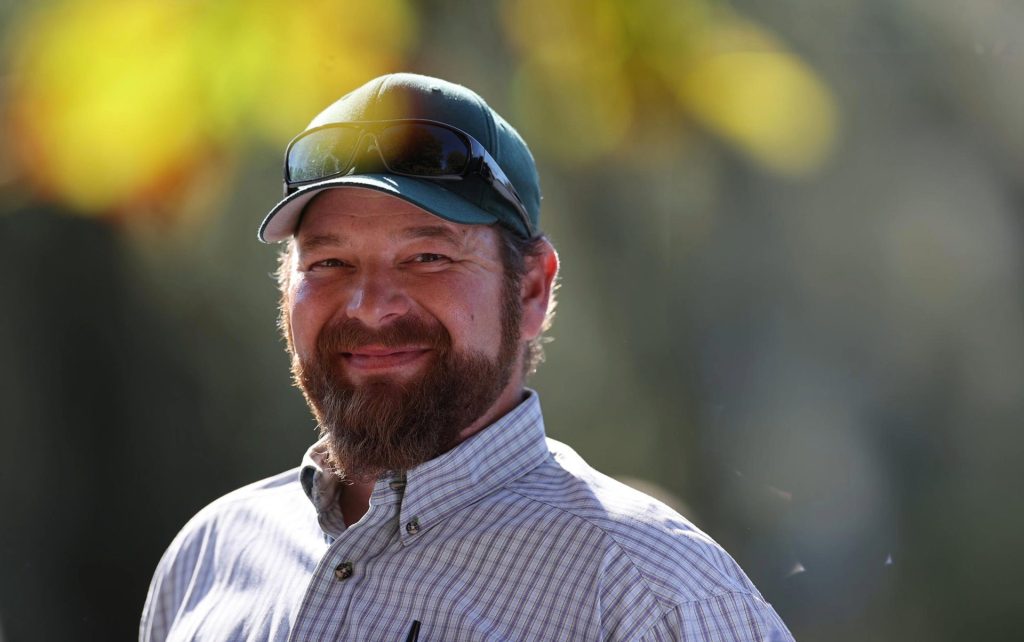 Porträt eines lächelnden Mannes mit Bart, der eine Kappe mit Sonnenbrille und ein kariertes Hemd trägt, vor einem unscharfen, sonnendurchfluteten Hintergrund.