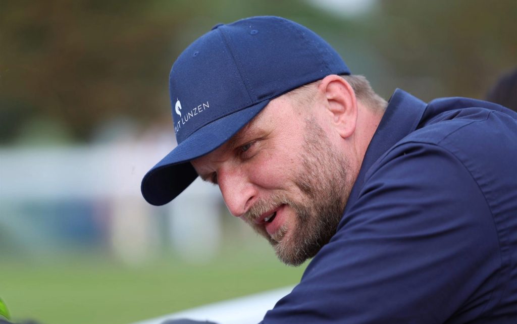 Mann mit Bart trägt eine dunkelblaue Kappe und ein dunkelblaues Hemd, Blick nach unten gerichtet.