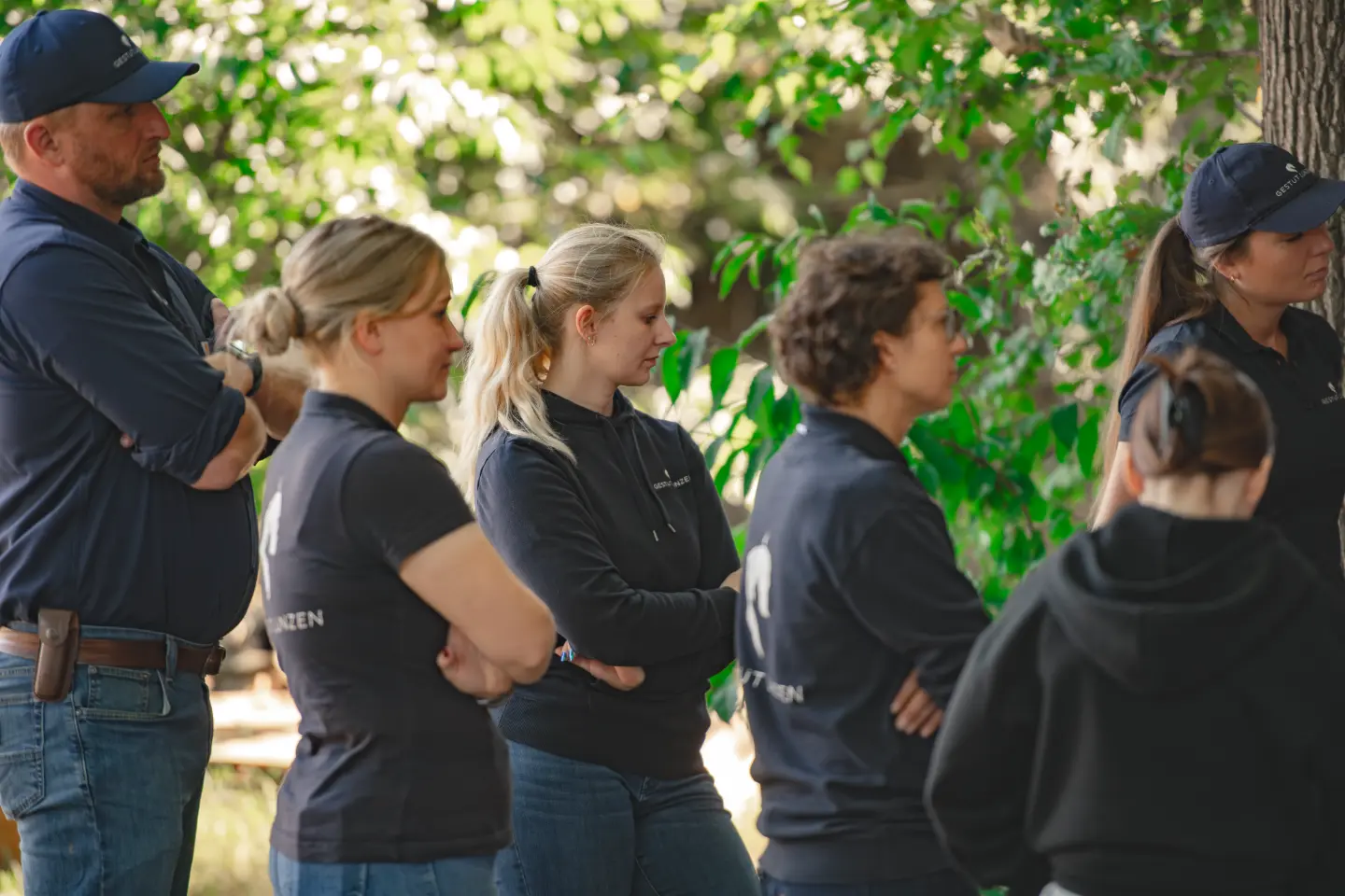 Fünf Personen in dunkler Freizeitkleidung stehen mit verschränkten Armen unter grünen Bäumen im Freien.