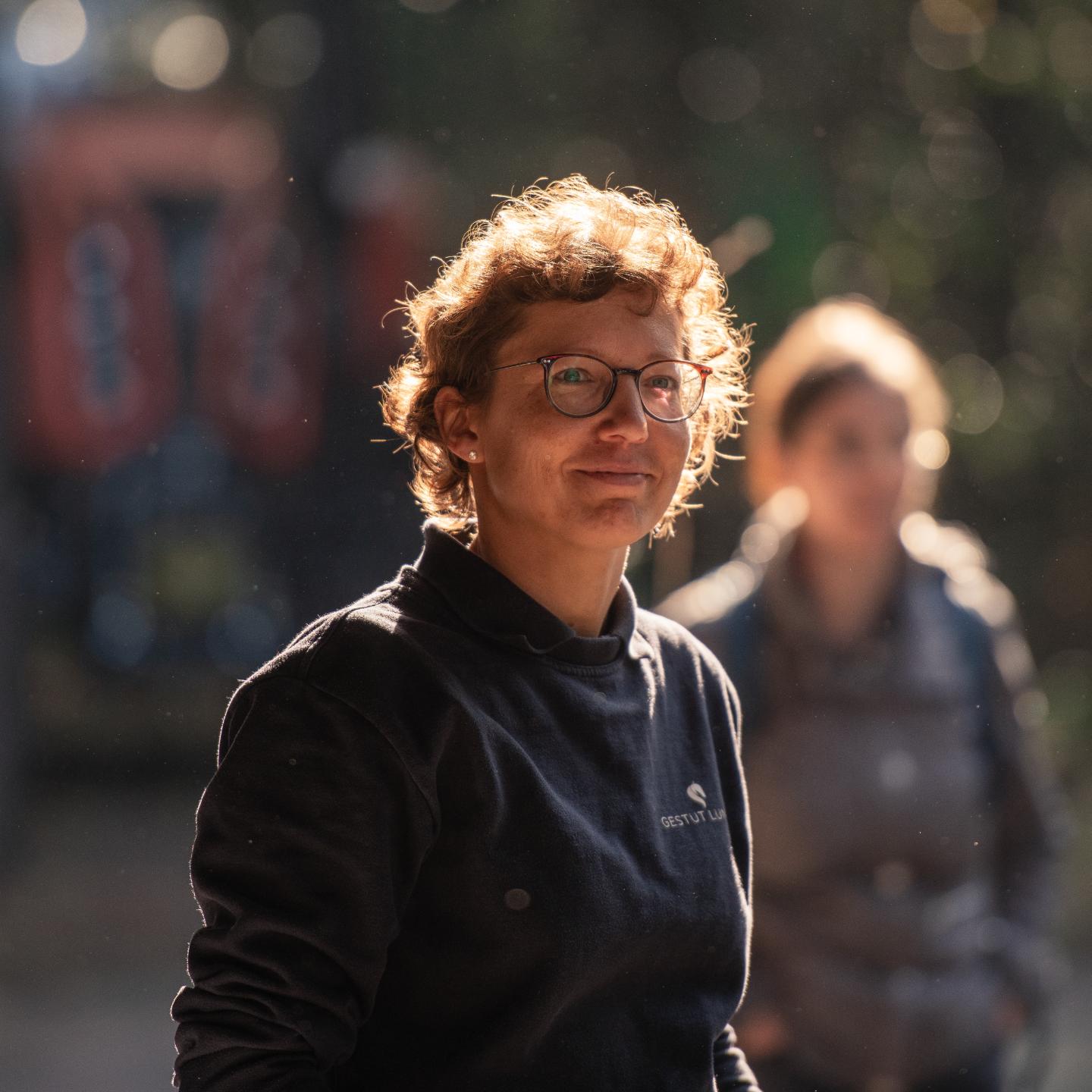 Frau mit kurzen lockigen Haaren und Brille, die bei Sonnenlicht in einem Outdoor-Umfeld lächelt.