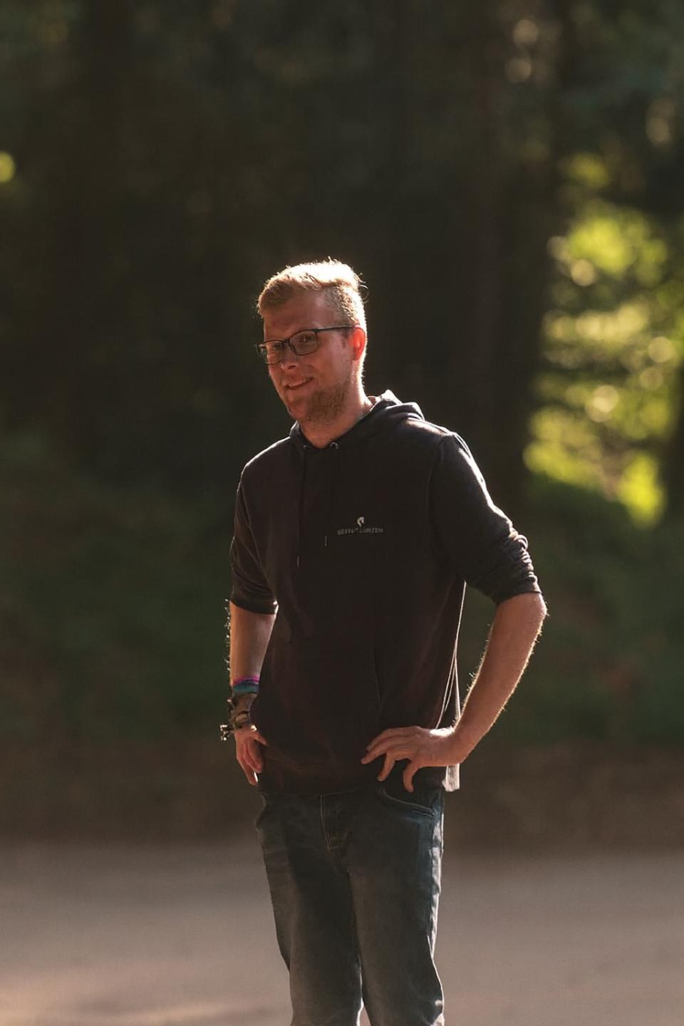 Mann mit Brille und Kapuzenpullover steht lächelnd draußen mit den Händen in den Hüften, Sonnenlicht fällt auf ihn.