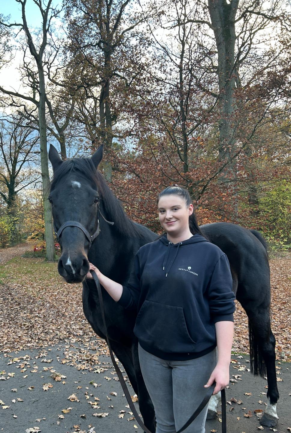 Junge Frau in schwarzem Kapuzenpullover hält ein schwarzes Pferd an der Führleine im herbstlichen Wald.