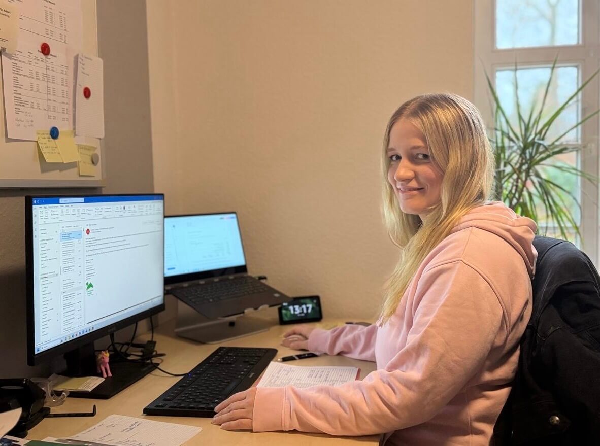 Junge Frau mit rosa Kapuzenpullover sitzt an einem Schreibtisch mit zwei Bildschirmen und lächelt zur Kamera.