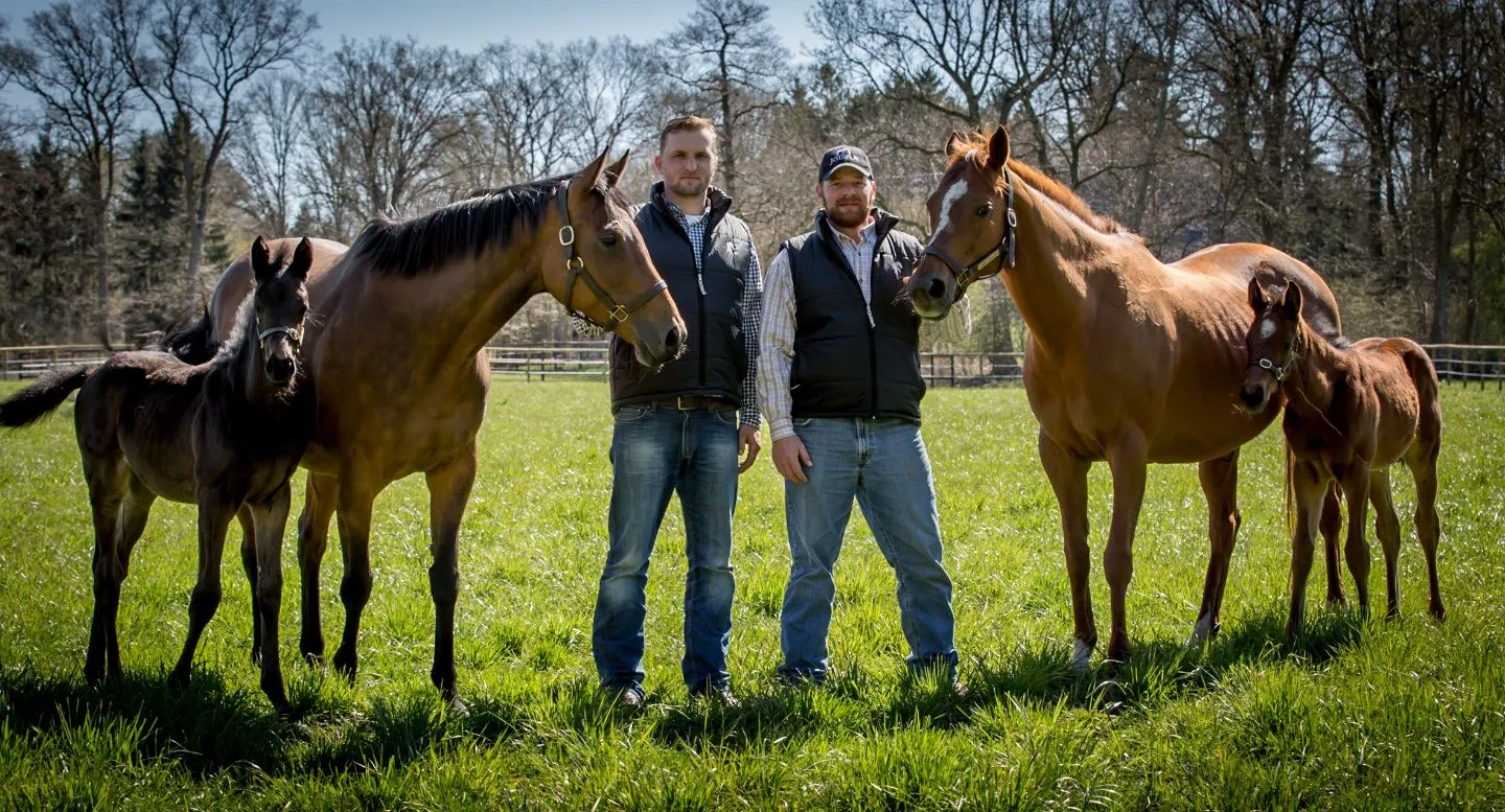 Zwei Männer in Weste und Jeans stehen auf einer grünen Weide mit jeweils einem erwachsenen Pferd und einem Fohlen.