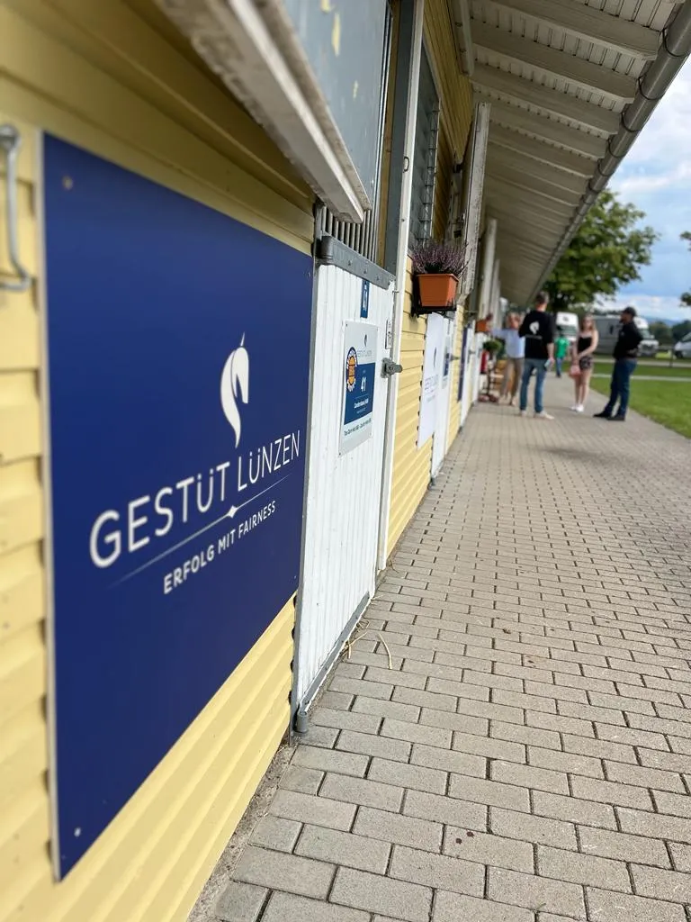 Blauer Schild mit der Aufschrift 'Gestüt Lünzen Erfolg mit Fairness' an gelber Stallwand, im Hintergrund mehrere Personen auf gepflastertem Weg.