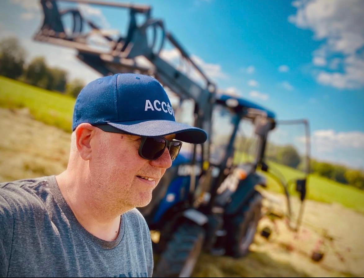 Mann mit Sonnenbrille und blauer Kappe steht auf einem Feld vor einem Traktor bei sonnigem Wetter.