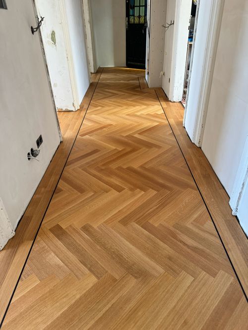 Langer Flur mit frisch verlegtem Fischgrät-Parkett von Damian Parkett & Boden, das den Gang optisch streckt und zur Tür am Ende führt.