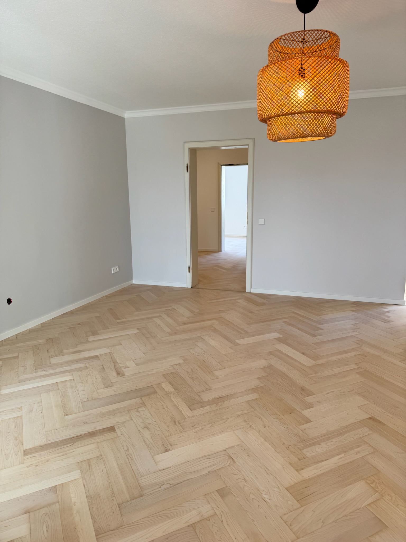 Leerer Altbauraum mit frisch verlegtem Fischgrät-Parkett von Damian Parkett & Boden, beleuchtet von einer modernen orangefarbenen Deckenlampe.