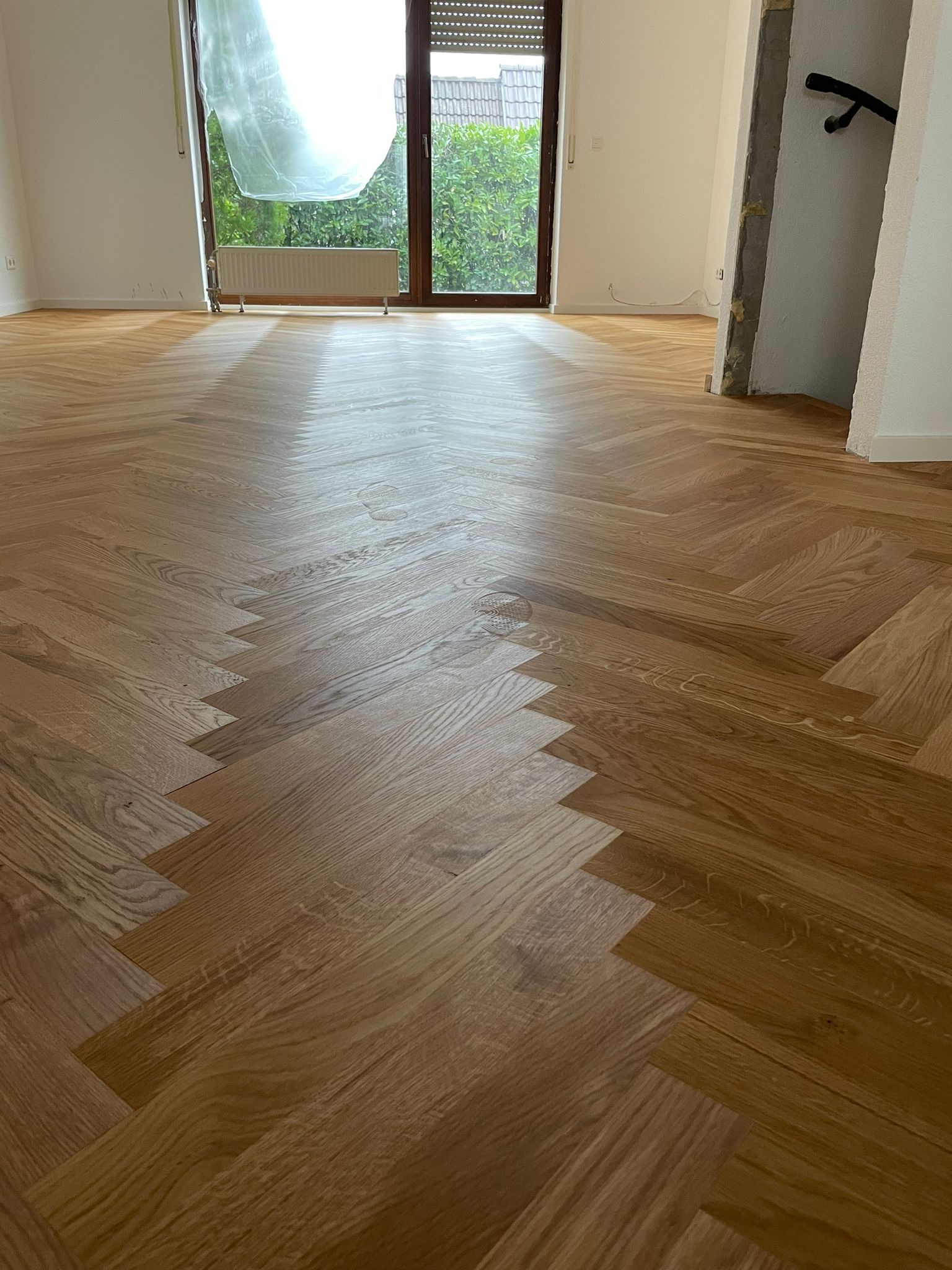 Neu verlegtes Fischgrät-Parkett von Damian Parkett & Boden, das zu einer Balkon- oder Terrassentür führt und den Blick ins Grüne freigibt.