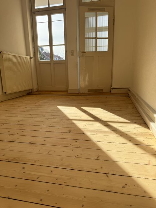 Leerer Altbauraum mit hellen, breiten Landhausdielen von Damian Parkett & Boden, auf die durch die Fensterfront starkes Sonnenlicht fällt.