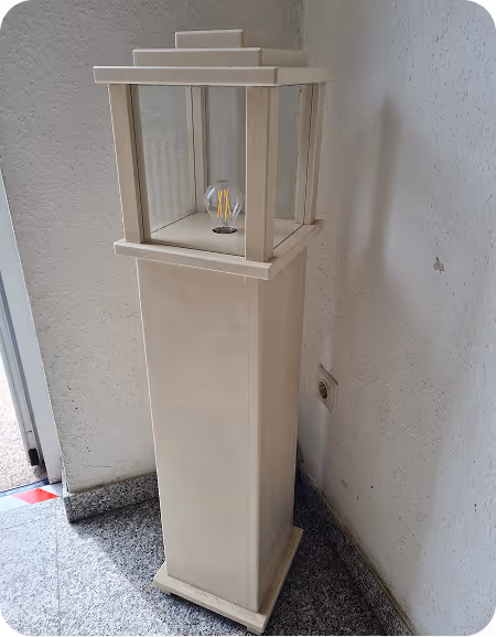 Ein beige-farbener Stehlampenständer mit einer sichtbaren Glühbirne, platziert in einer Ecke vor einer weißen Wand in einem Raum mit grauem Boden.