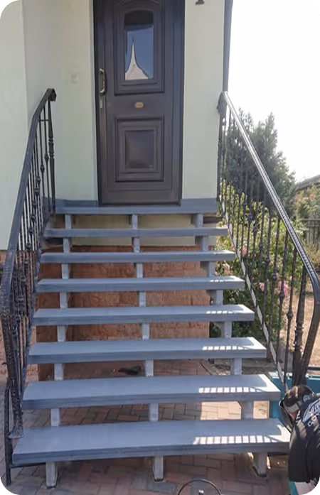 Außentreppe mit acht grauen Stufen und schwarzen Metallgeländern führt zu einer dunklen Haustür an einem hellen Gebäude.