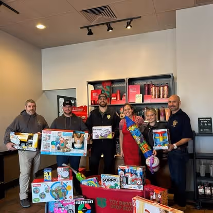 Five adults standing indoors holding various toy boxes with more toys stacked on the floor in front of them for a toy drive event.