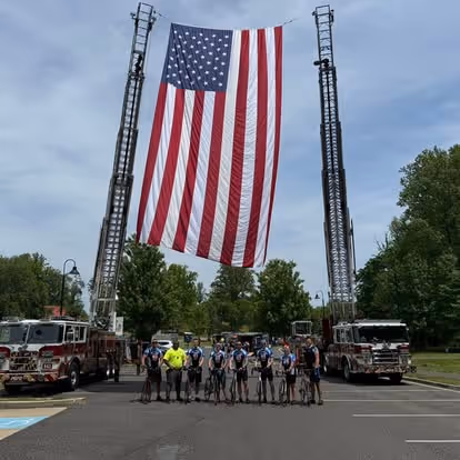 Large American flag hanging between two extended fire truck ladders with a group of cyclists standing beneath on a street.