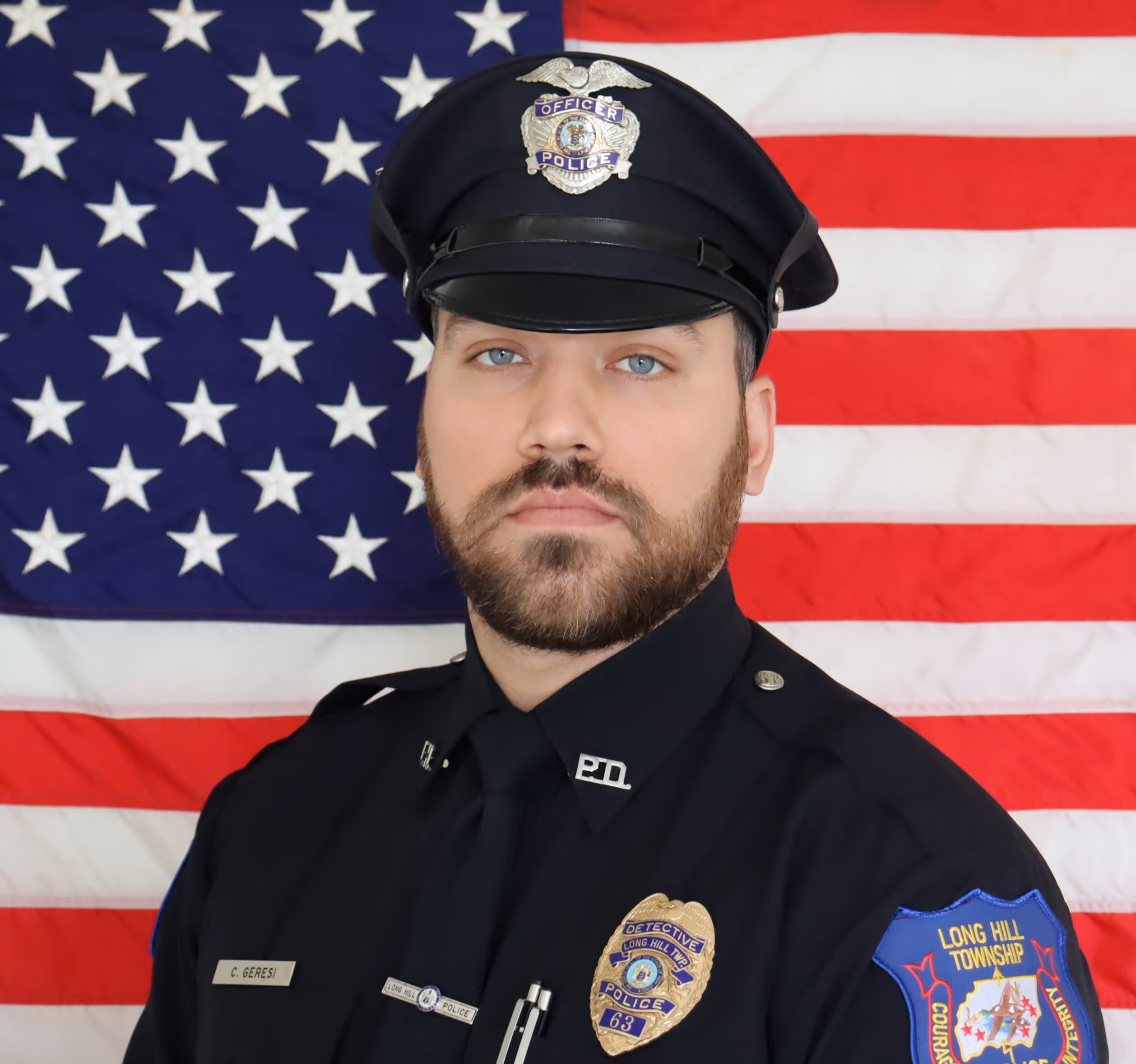 Police officer in uniform with badge and name tag standing in front of an American flag.