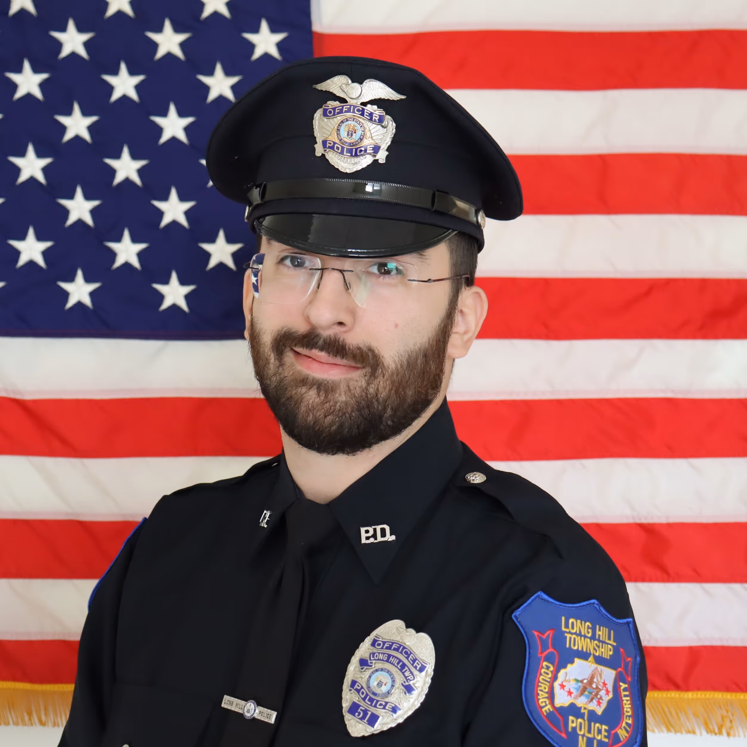 Portrait of a police officer in uniform with glasses and beard, standing in front of an American flag.