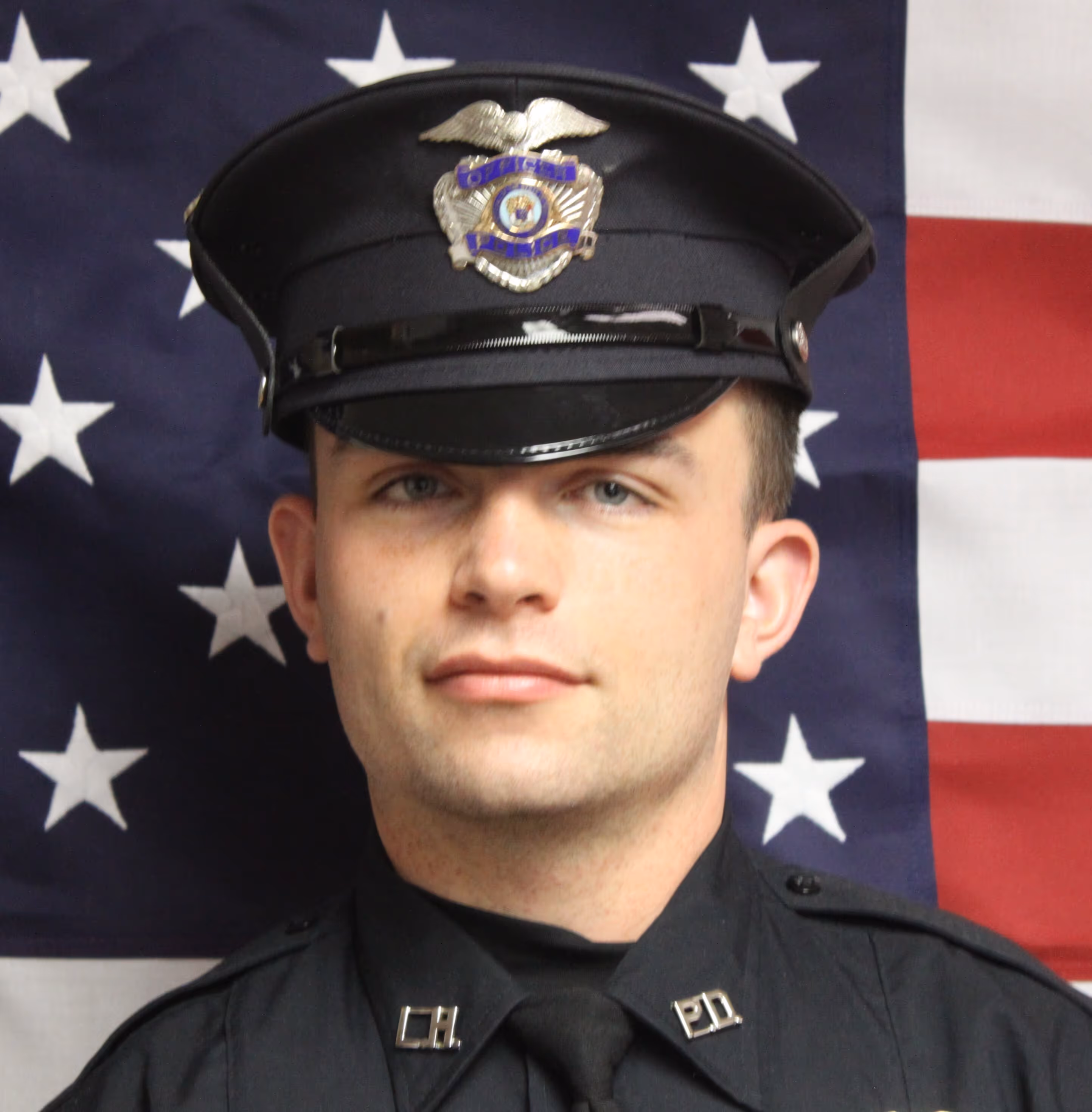 Portrait of a male police officer in uniform and cap standing in front of an American flag backdrop.
