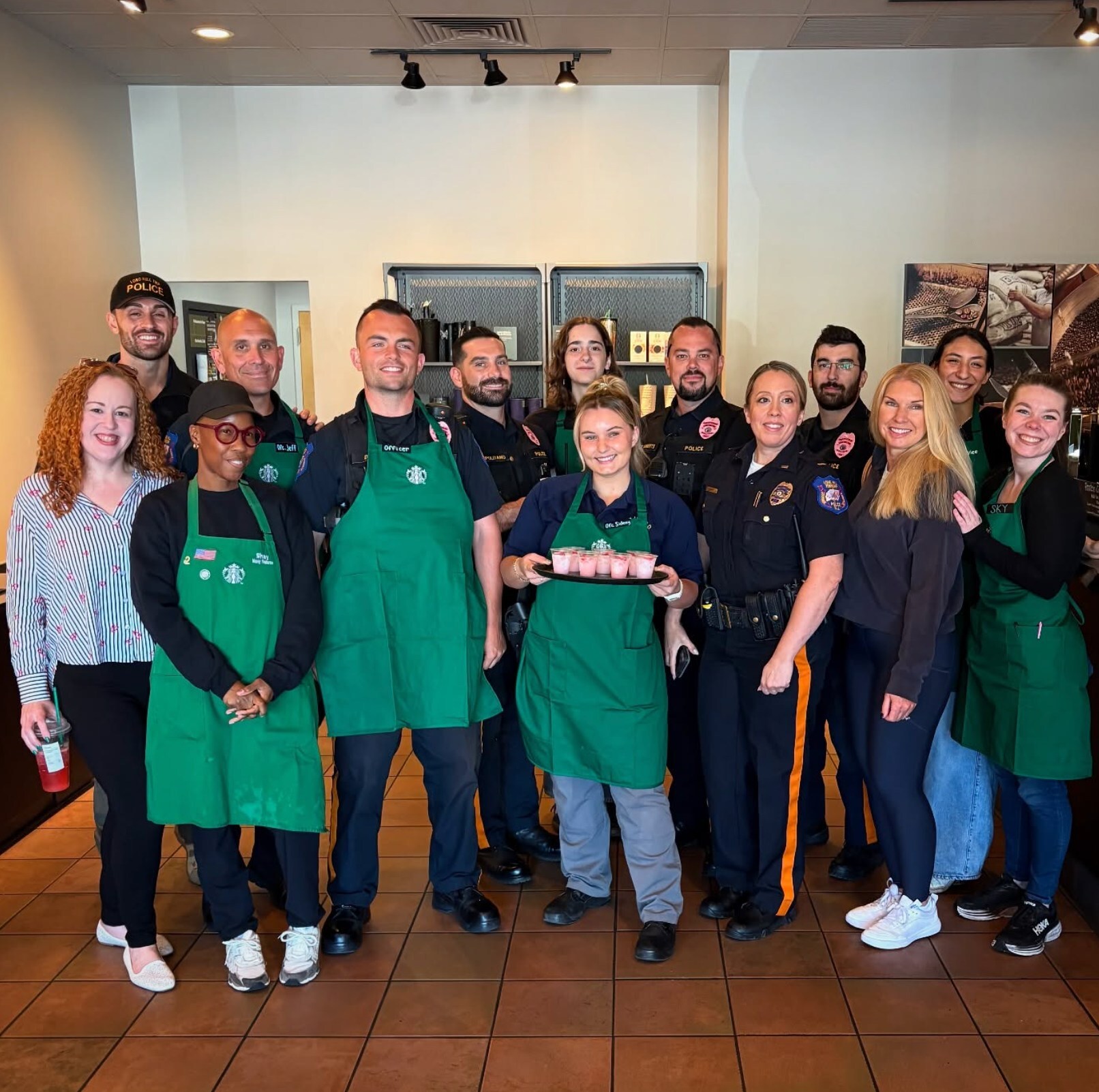 Group of Starbucks employees and police officers smiling inside a Starbucks store, with one employee holding a tray of pink drink samples.